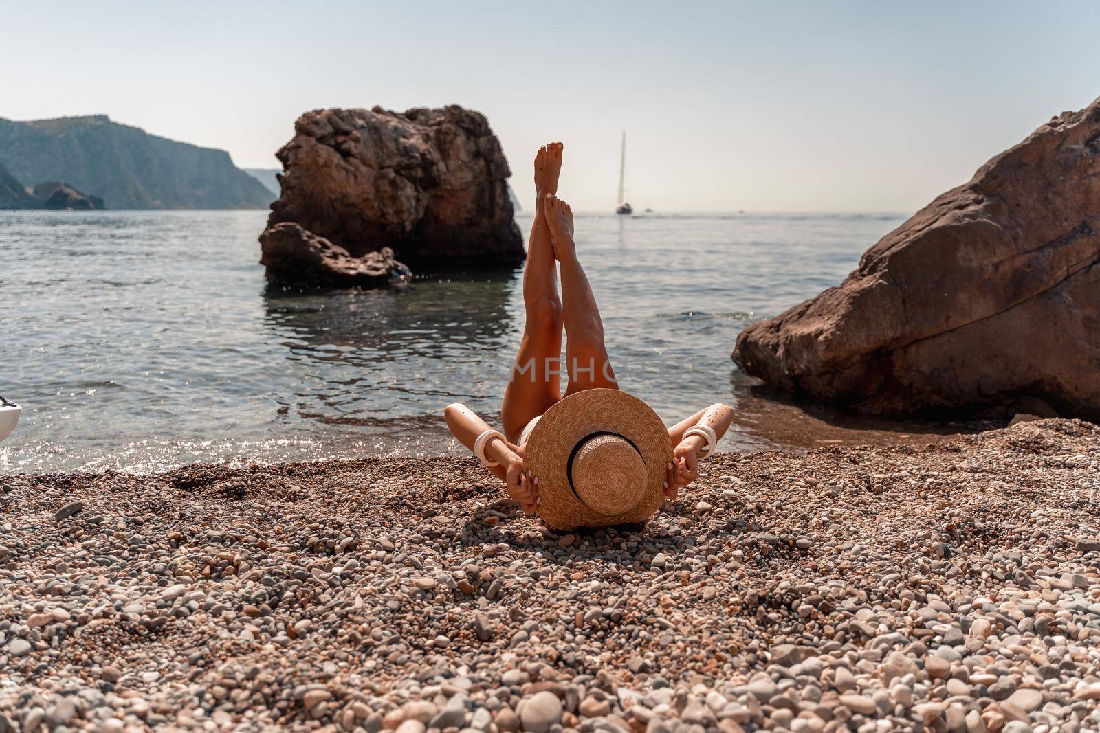 A beautiful middle-aged woman lies on the beach with her feet to the sky, covering her body with a wide-brimmed straw hat. She is sunbathing on the ocean. Vacation, travel, vacation concept. by Matiunina