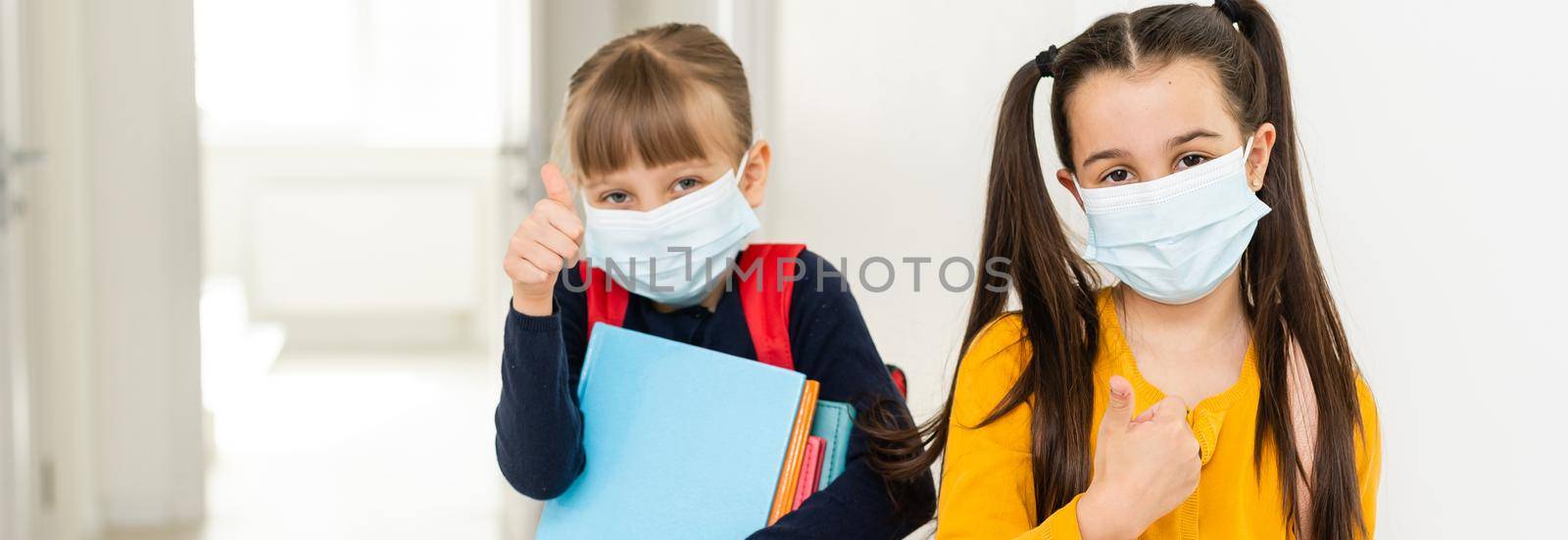 Cute little girls of primary hug each other with the mask on. School girls carry notebooks in hand and greet each other before entering school. by Andelov13