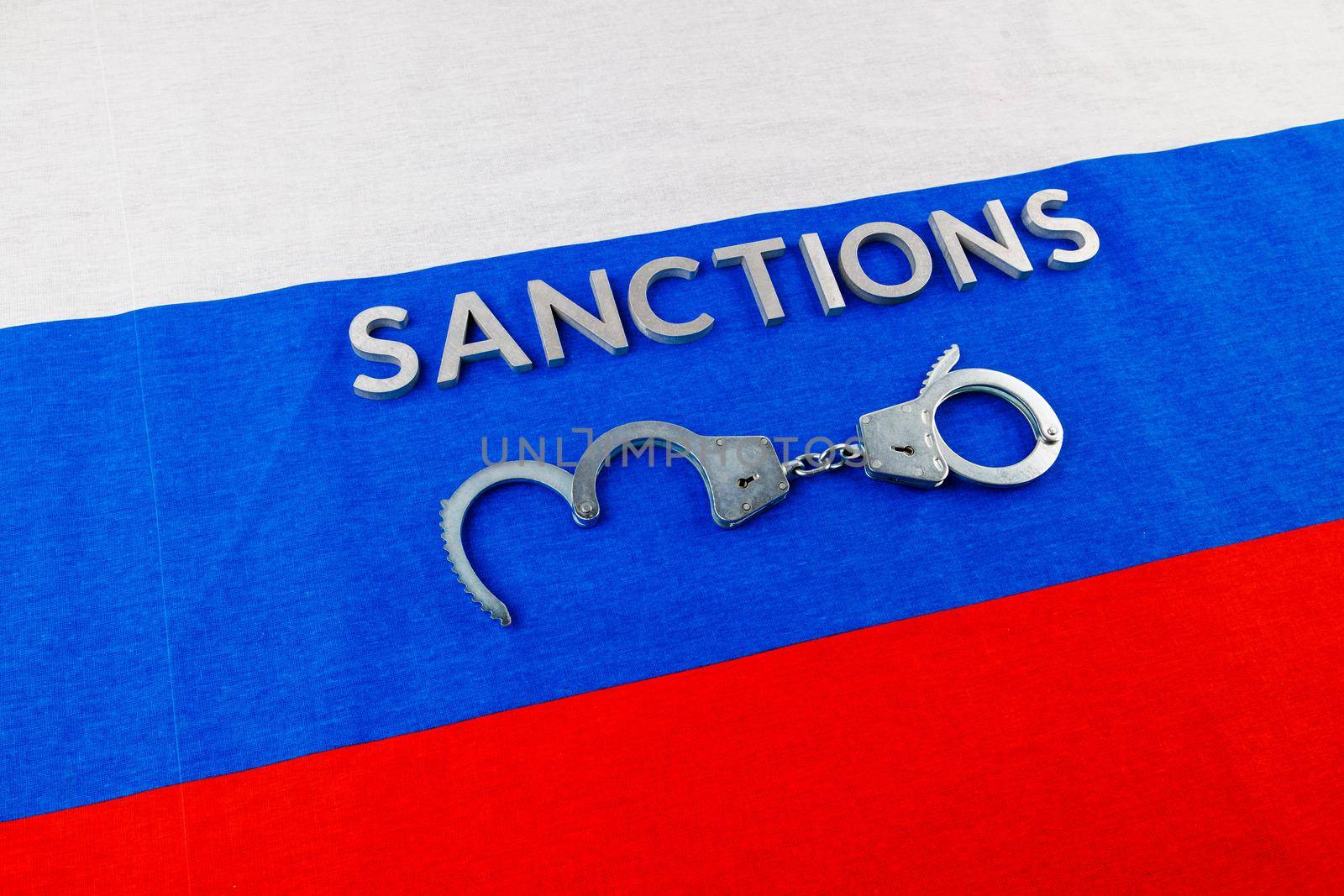 the word sanctions laid with silver metal letters on russian tricolor flag near handcuffs over it in directly above view