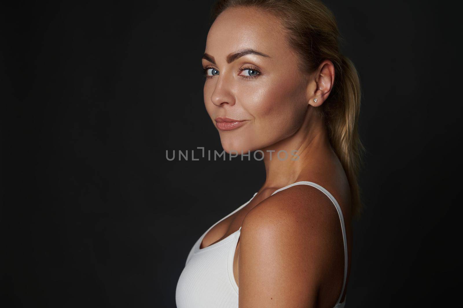 Close-up portrait of a beautiful slim body Caucasian woman, fit and muscular build young fitness girl in white top confidently looking at camera, isolated over black background with copy ad space by artgf