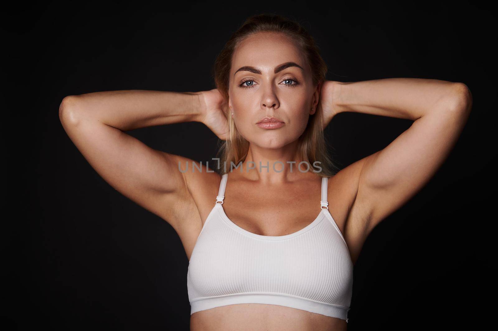 Beauty portrait of an attractive confident blonde European woman in white underclothing holding her hands behind head, looks at camera posing against black background with copy ad space