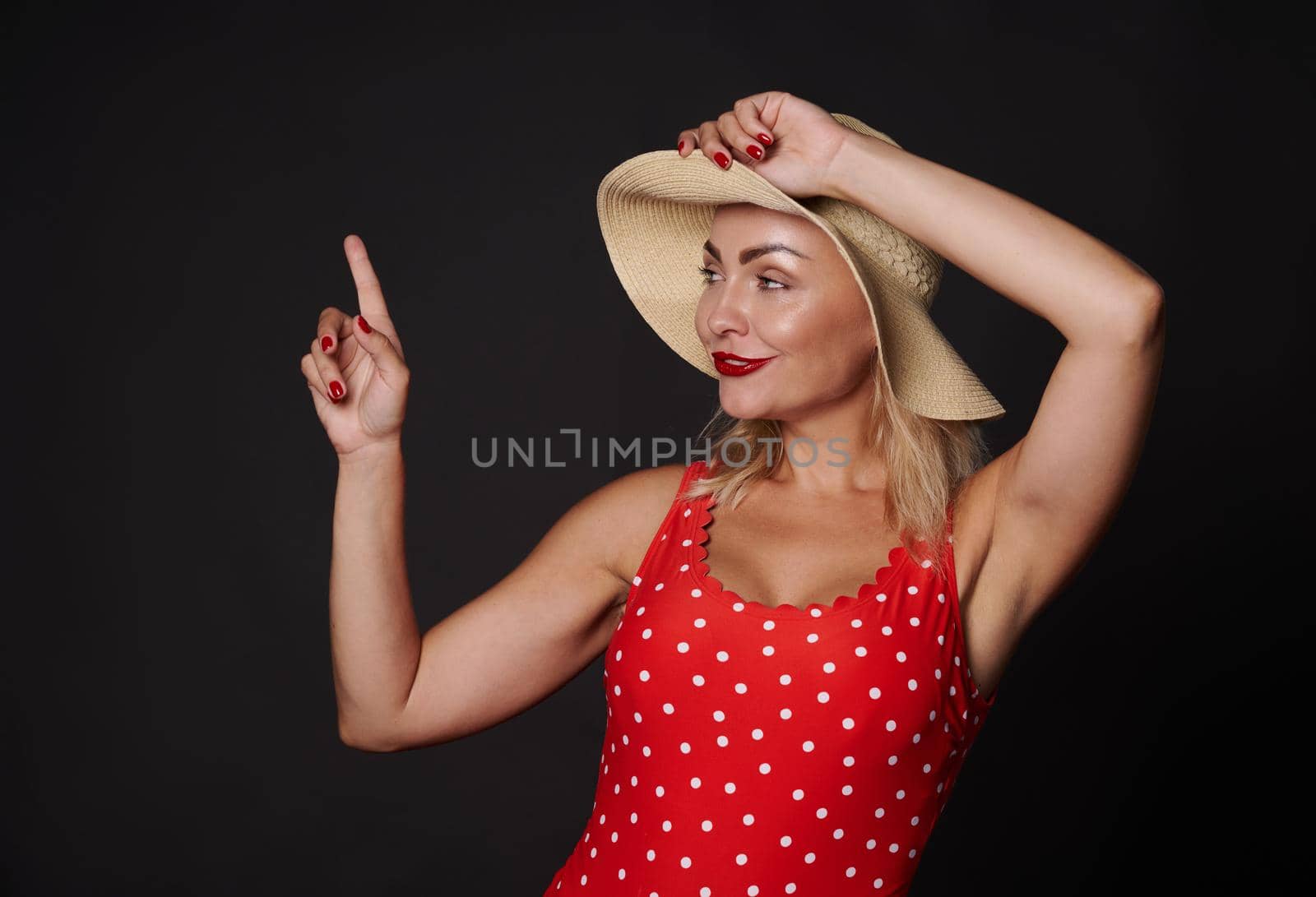 Cheerful attractive adorable blonde European woman in red swimsuit with white polka dots and straw summer hat pointing her finger up at copy space on black background. Summer advertising concept