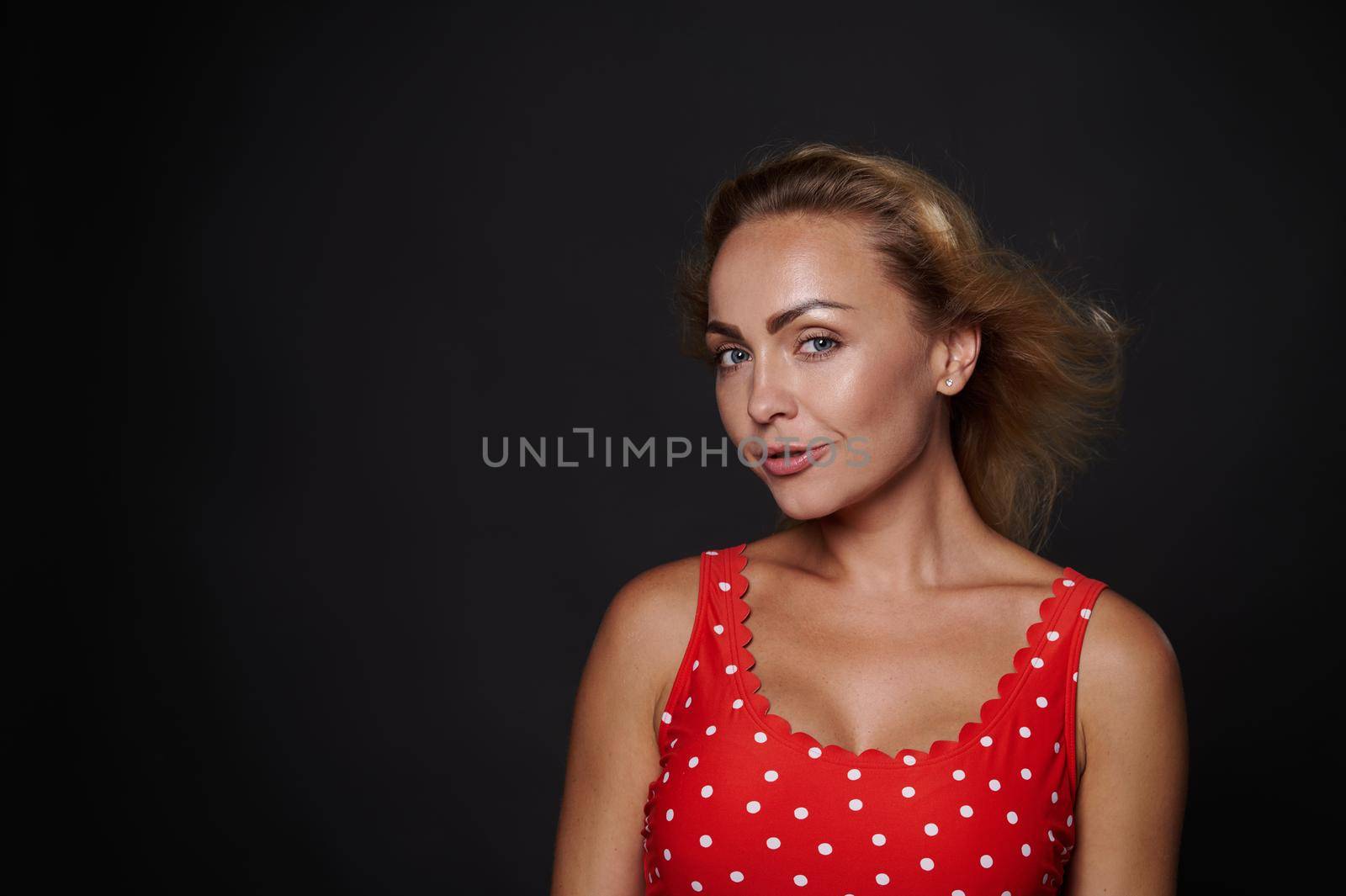 Close-up beauty portrait of an attractive gorgeous middle aged European woman in red top with white polka dots confidently looking at camera isolated over black background with copy ad space