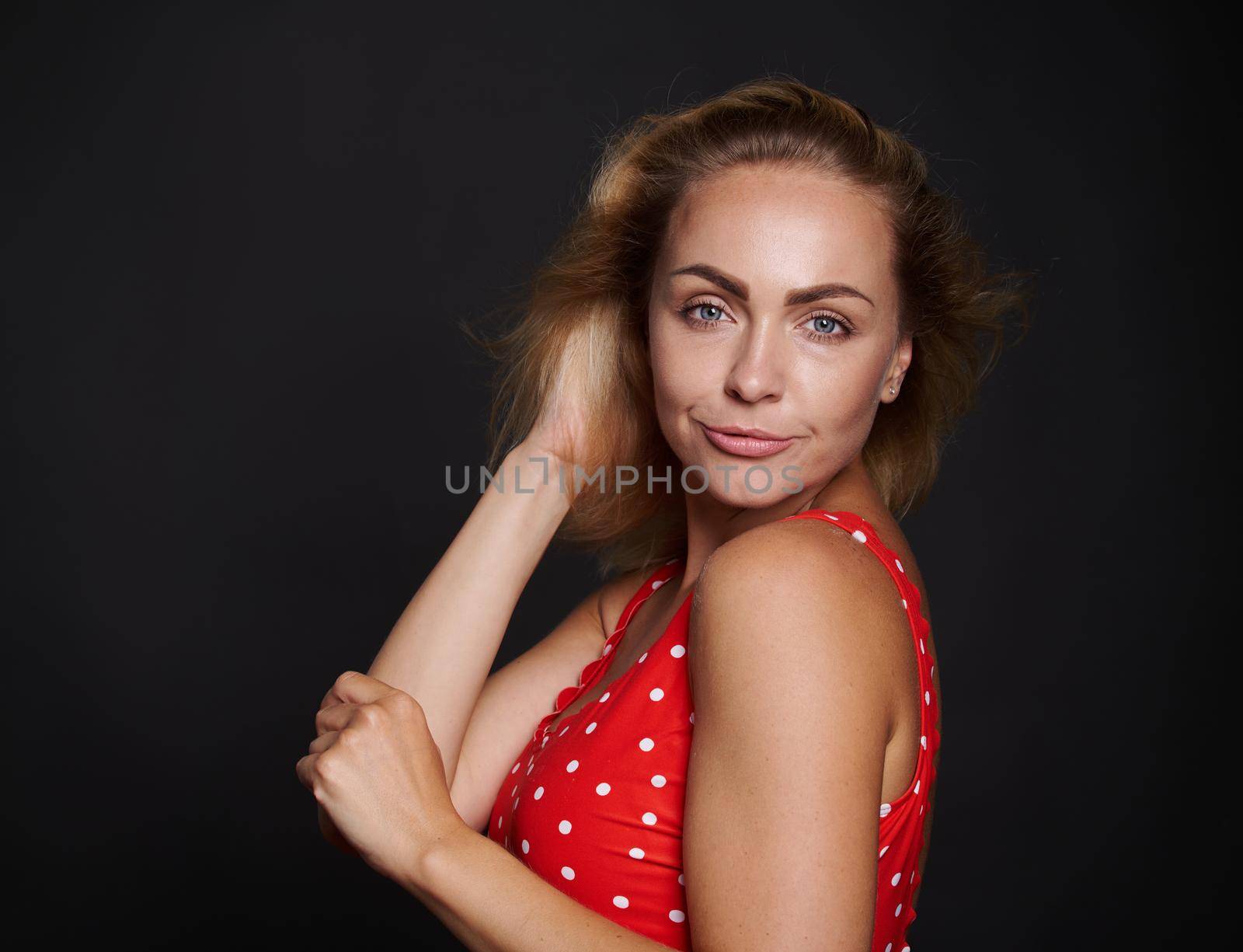 Beautiful middle aged blonde sporty European woman in red swimsuit with white polka dots confidently looking at camera isolated over black background with copy ad space by artgf