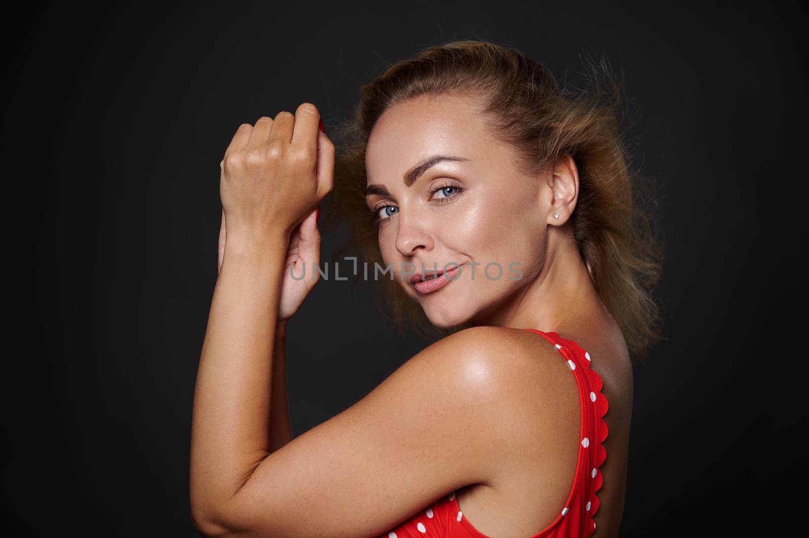 Portrait of a beautiful middle aged Caucasian woman with natural makeup and healthy glowing tanned skin, shiny flying blond hair, wearing red swimsuit isolated over black background with copy space by artgf