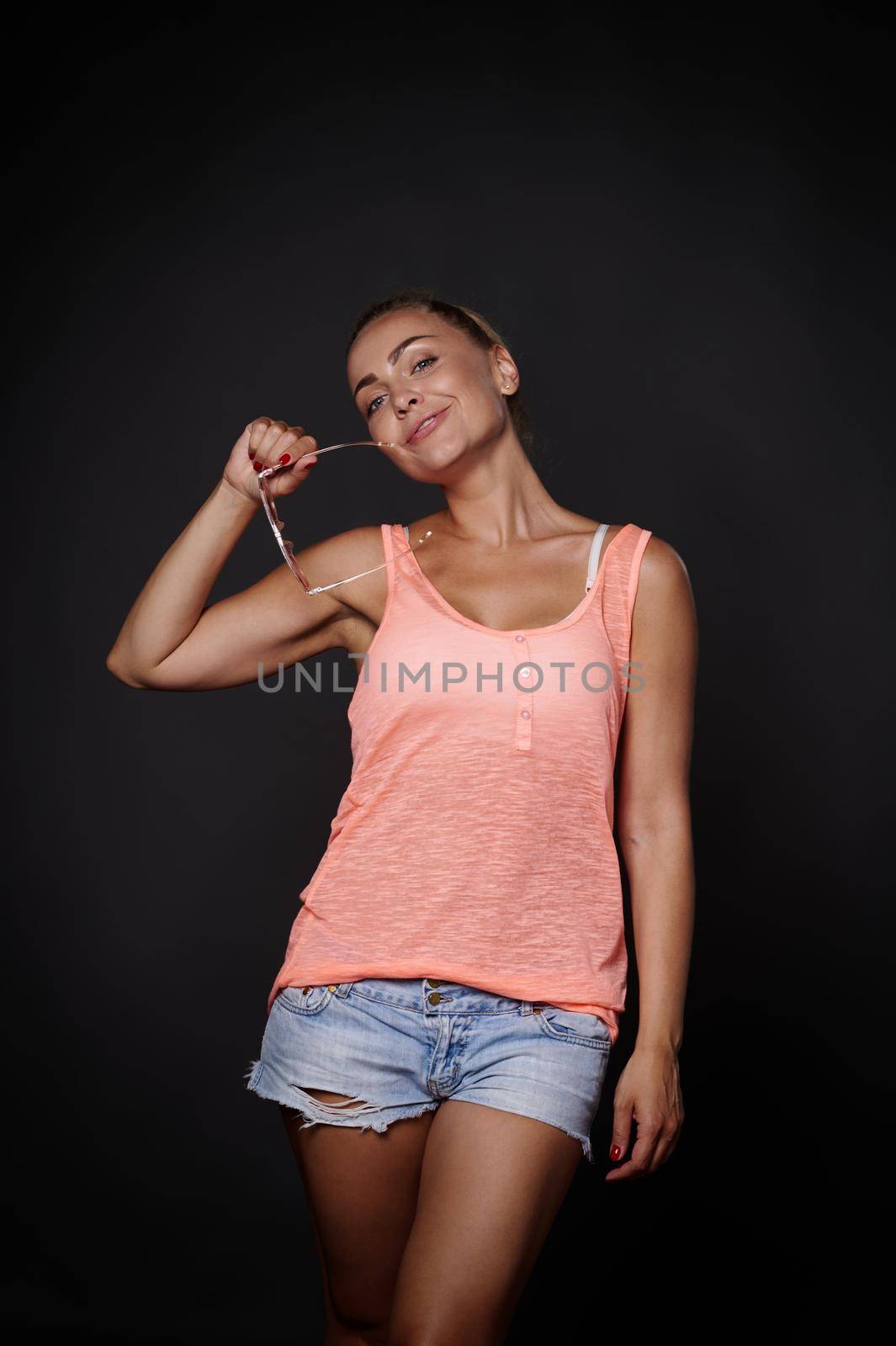 Charming sexy young blonde Caucasian pretty woman with tanned skin, aesthetic fit body in denim shorts, pink top and sunglasses posing over black background with copy ad space by artgf