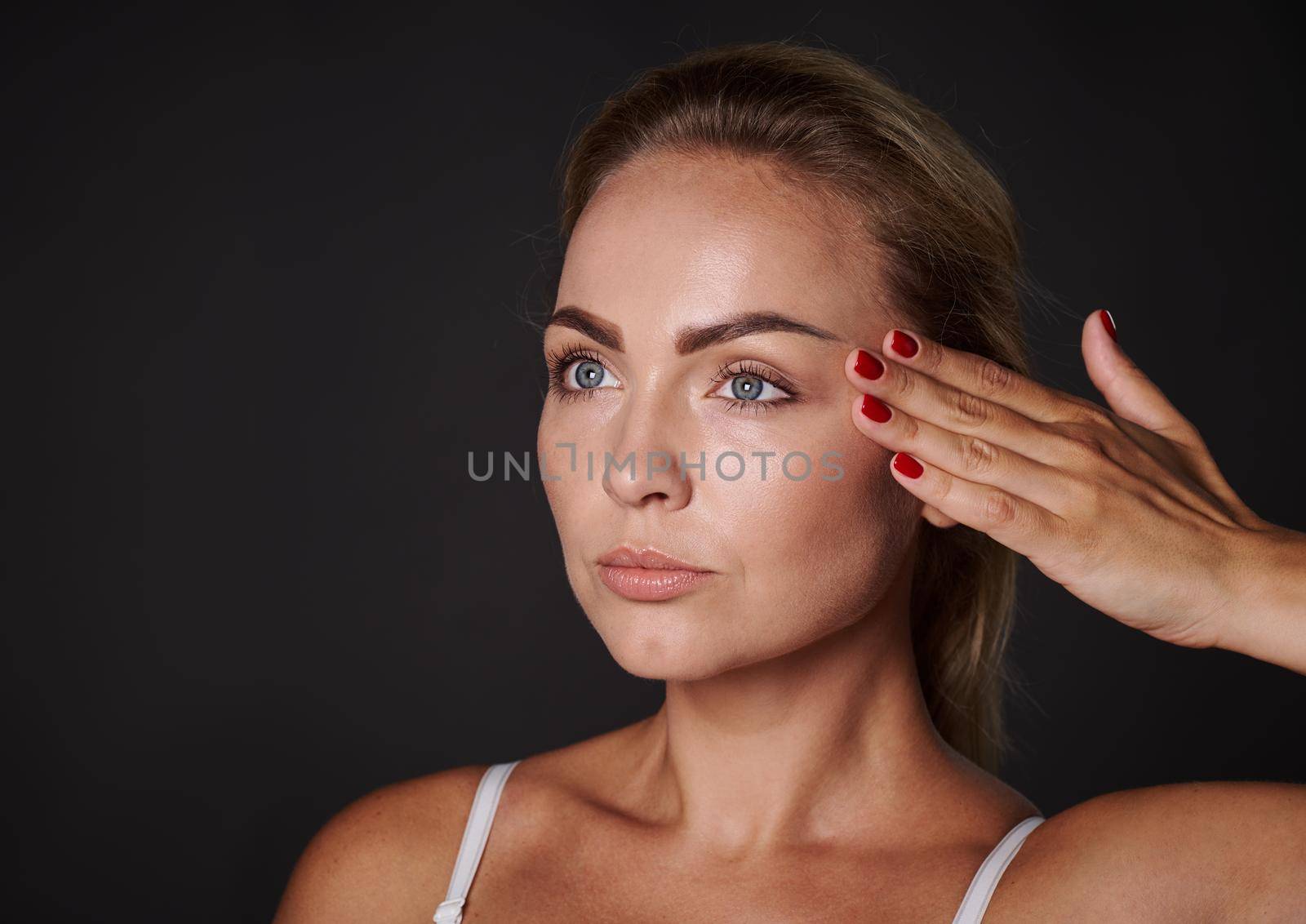 Close-up beauty portrait of a beautiful 30 years old blonde Caucasian woman with glowing fresh healthy skin holding her hand on her temple, isolated over black background with copy space