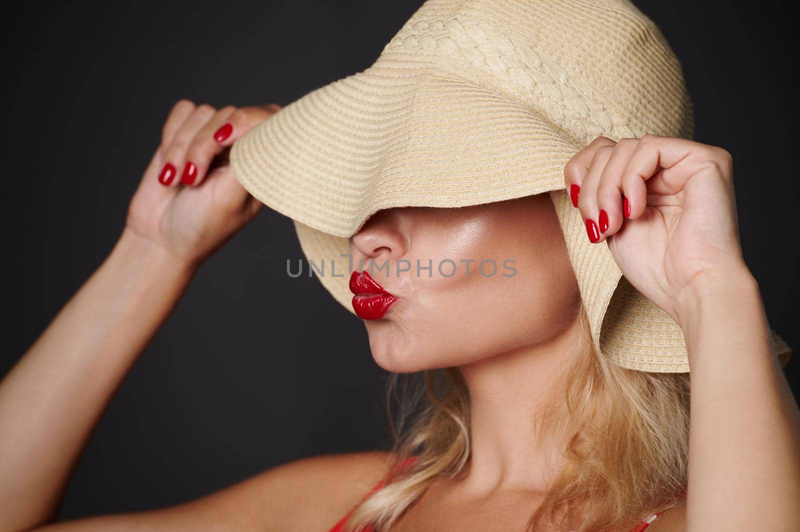 Close-up beautiful Caucasian woman covering her eyes with a straw summer hat and sending kiss, isolated over black background with copy space. Headshot portrait of a pretty lady with red lipstick by artgf