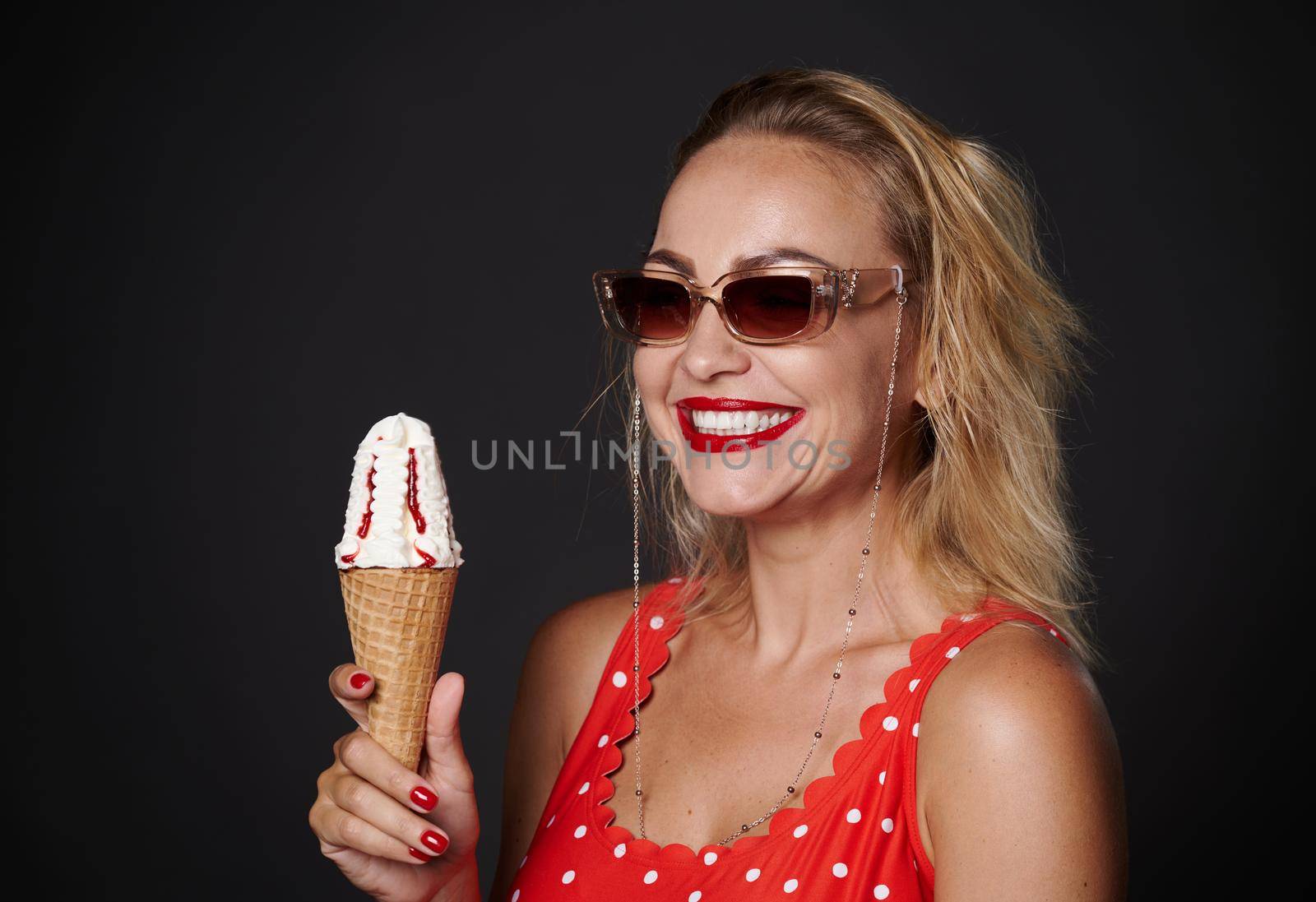 Cheerful middle aged blonde woman wearing protective sunglasses and red swimsuit, holding a ice-cream cone and smiles toothy smile looking at camera, isolated over black background with copy space by artgf