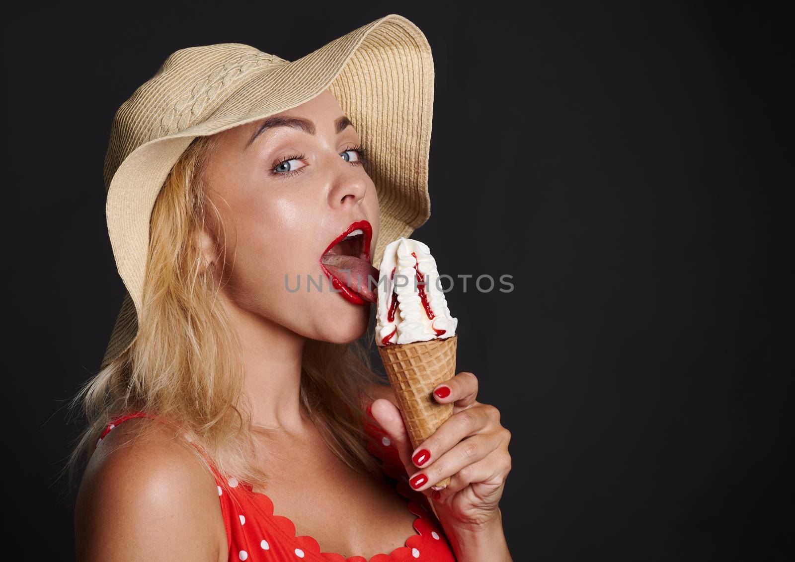 Headshot pretty blonde Caucasian woman in summer straw hat and red swimsuit with white polka dots tasting a delicious strawberry ice cream cone posing against black background with copy space for ads