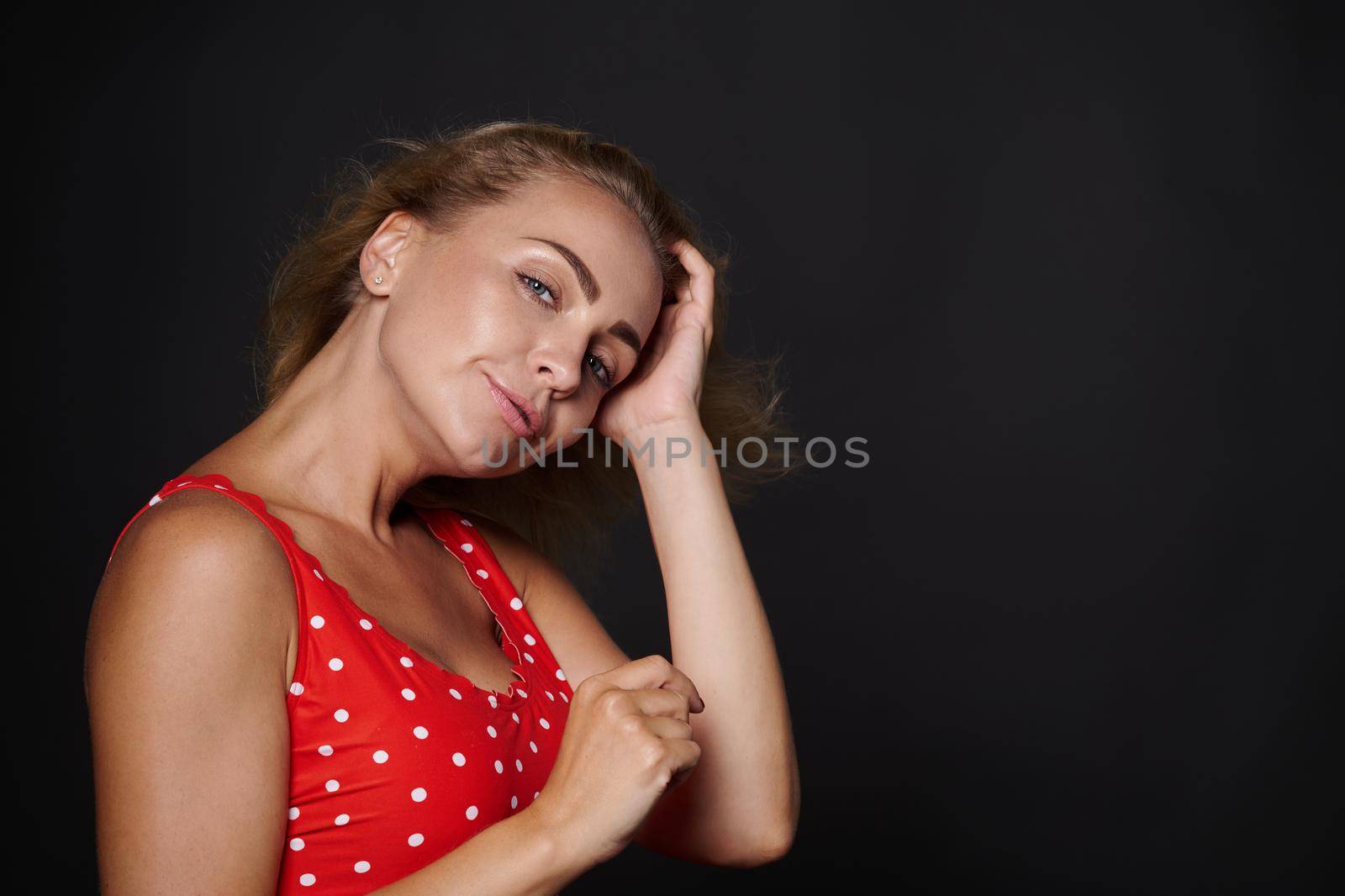 Beautiful Caucasian woman with healthy clean fresh glowing tanned skin and blond shiny straight hair, natural makeup, wearing red swimsuit with white polka dots isolated on black background