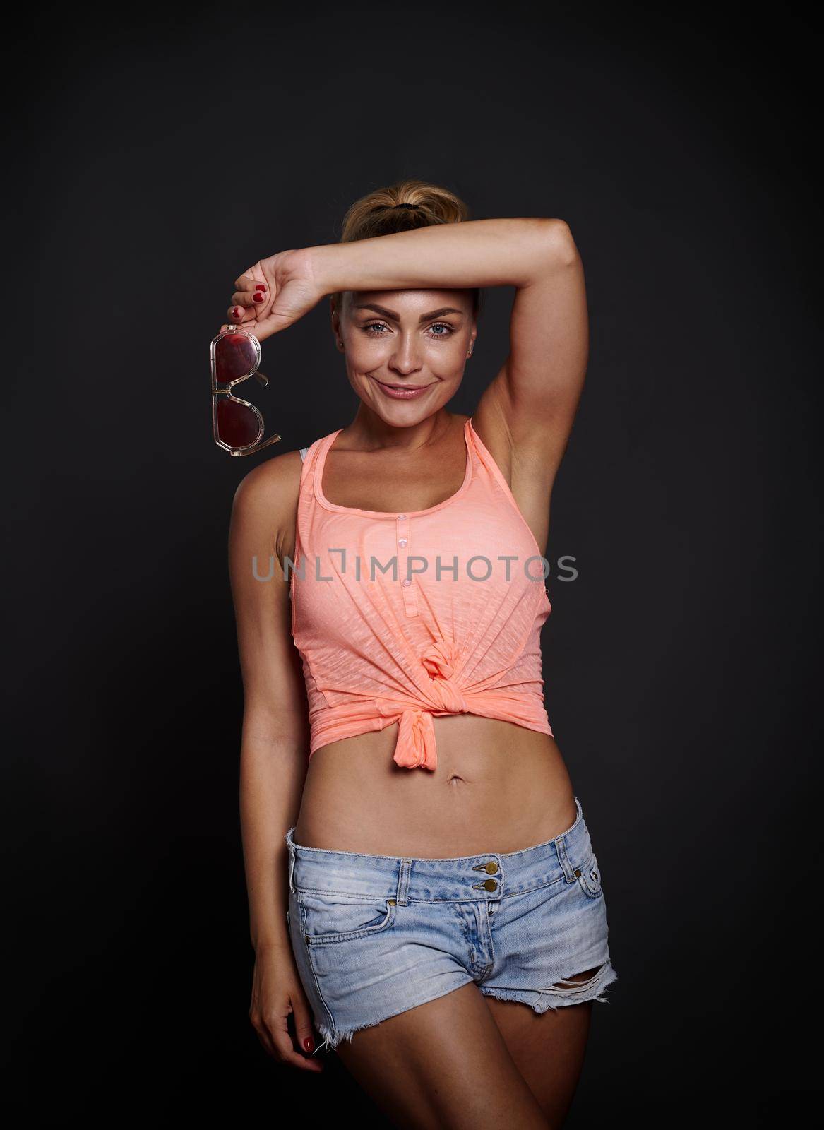 Fashion portrait of a stunning glamour blonde Caucasian woman in summer outfit, with beautiful aesthetic fit body and flat belly posing against black background with copy space by artgf