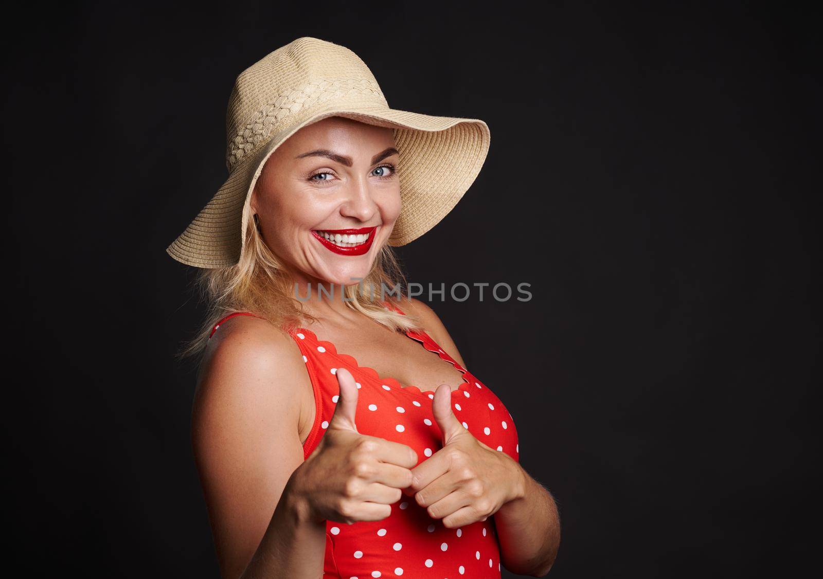 Attractive stunning blonde woman in summer straw hat and red swimsuit winking and smiling with beautiful white toothy smile, showing thumbs up at camera, posing over black background with copy space by artgf