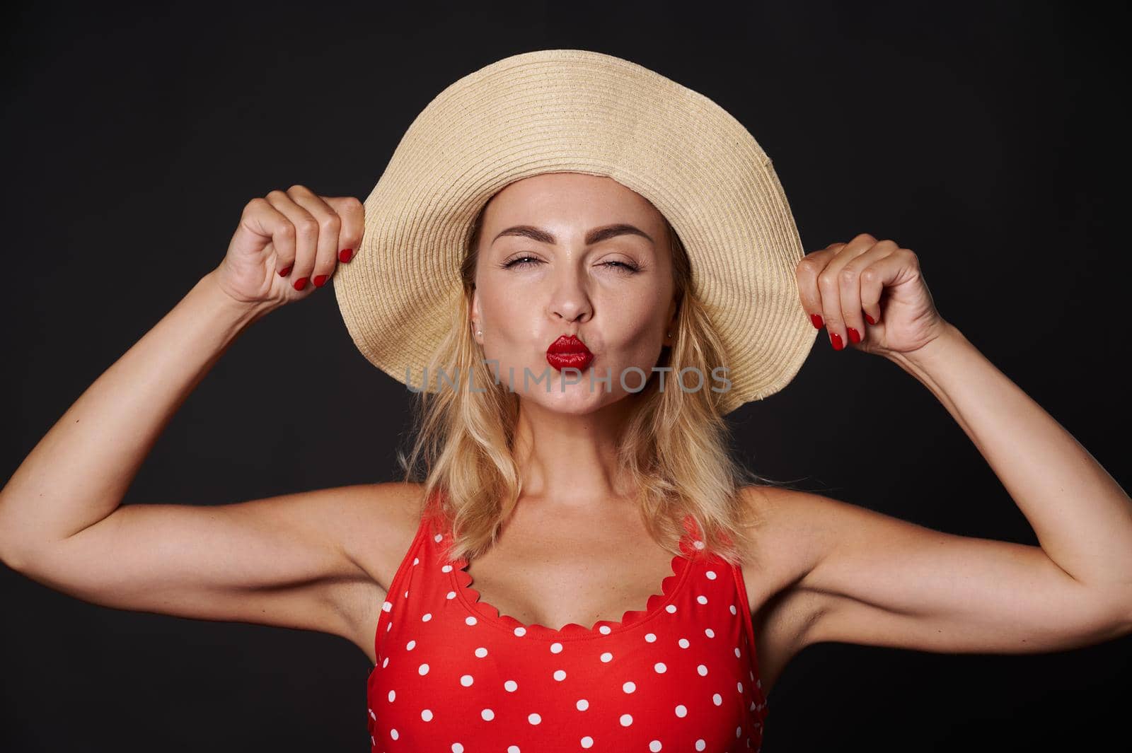 Close-up portrait of a beautiful sexy delightful stunning blonde European woman in red swimsuit with white polka dots and a straw summer hat sending a kiss, against black background with copy space by artgf