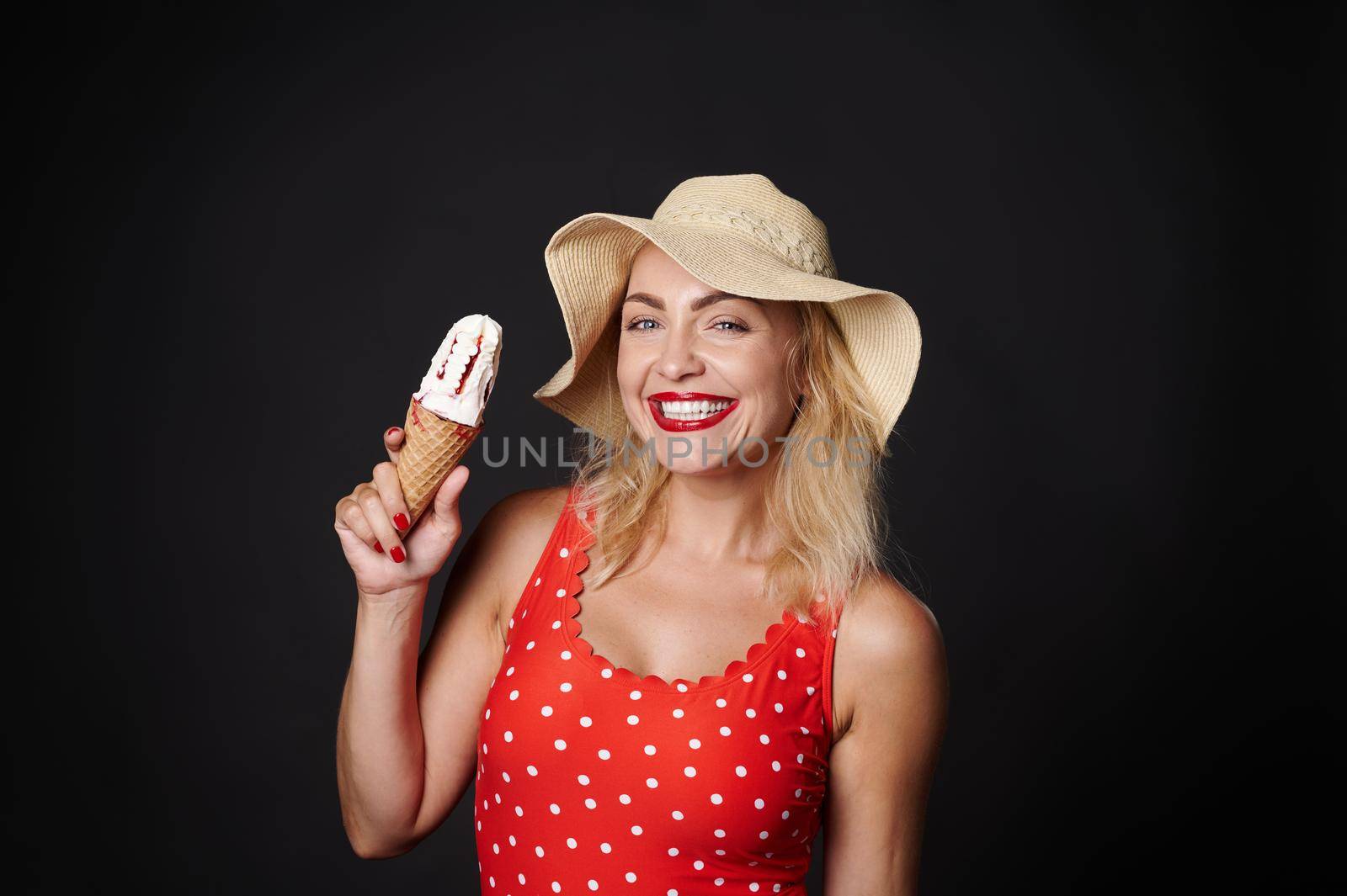 Beautiful Caucasian blonde woman in red swimsuit with white polka dots and summer straw hat with strawberry ice cream cone smiles with cheerful toothy smile against black background with copy ad space