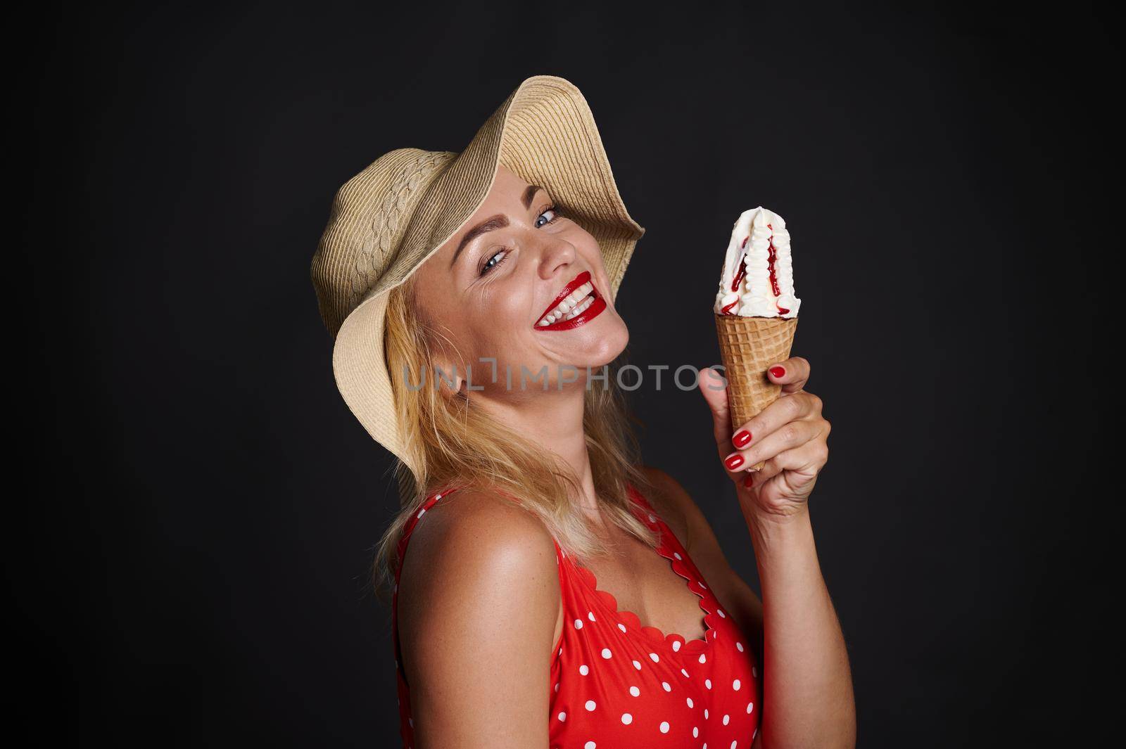 Gorgeous cheerful European woman in straw summer hat red swimsuit with white polka dots, holding delicious ice cream cone , smiling beautiful toothy smile, posing on black background copy space