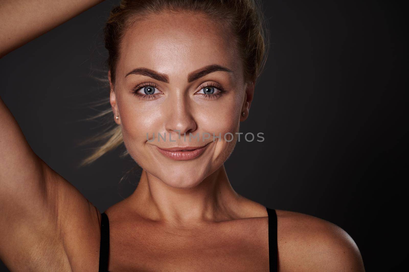 Close-up portrait of the face of a beautiful woman with fresh clean glowing tanned skin, smiling looking at camera isolated over dark background. Body positivity, self-acceptance, confidence concept