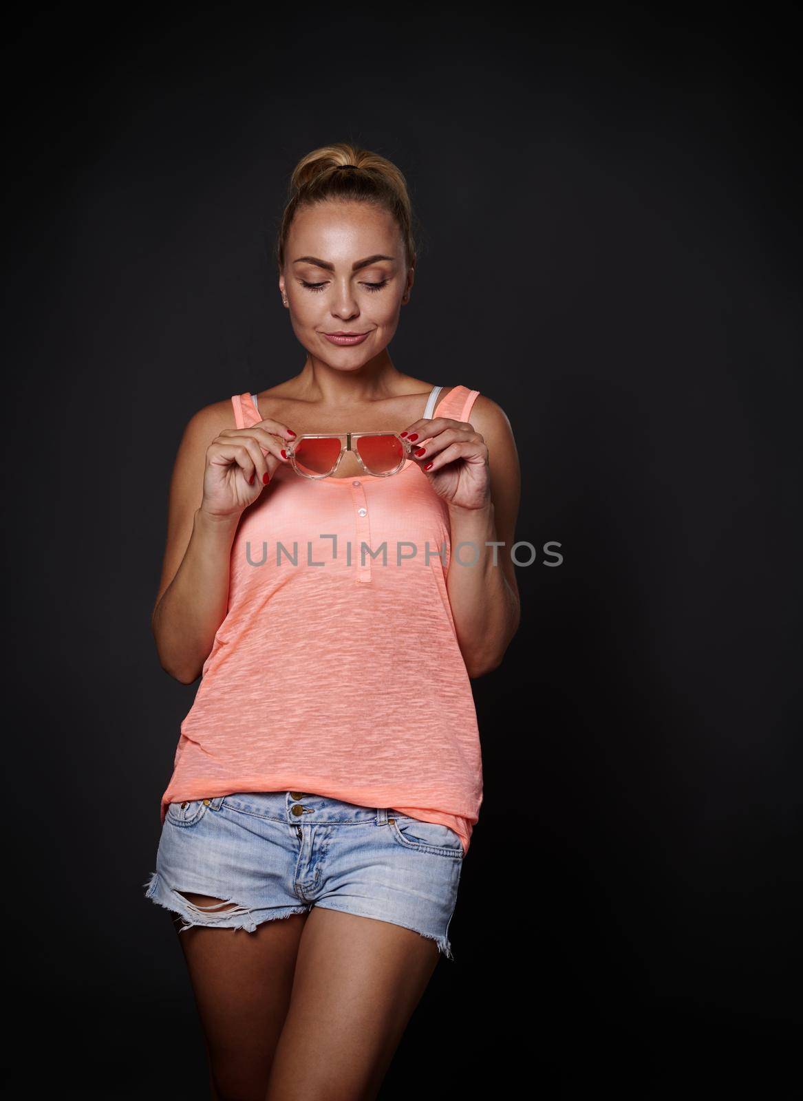 Charming sexy young blonde Caucasian pretty woman with tanned skin, aesthetic fit body in denim shorts, pink top holding sunglasses and smiling posing over black background with copy ad space