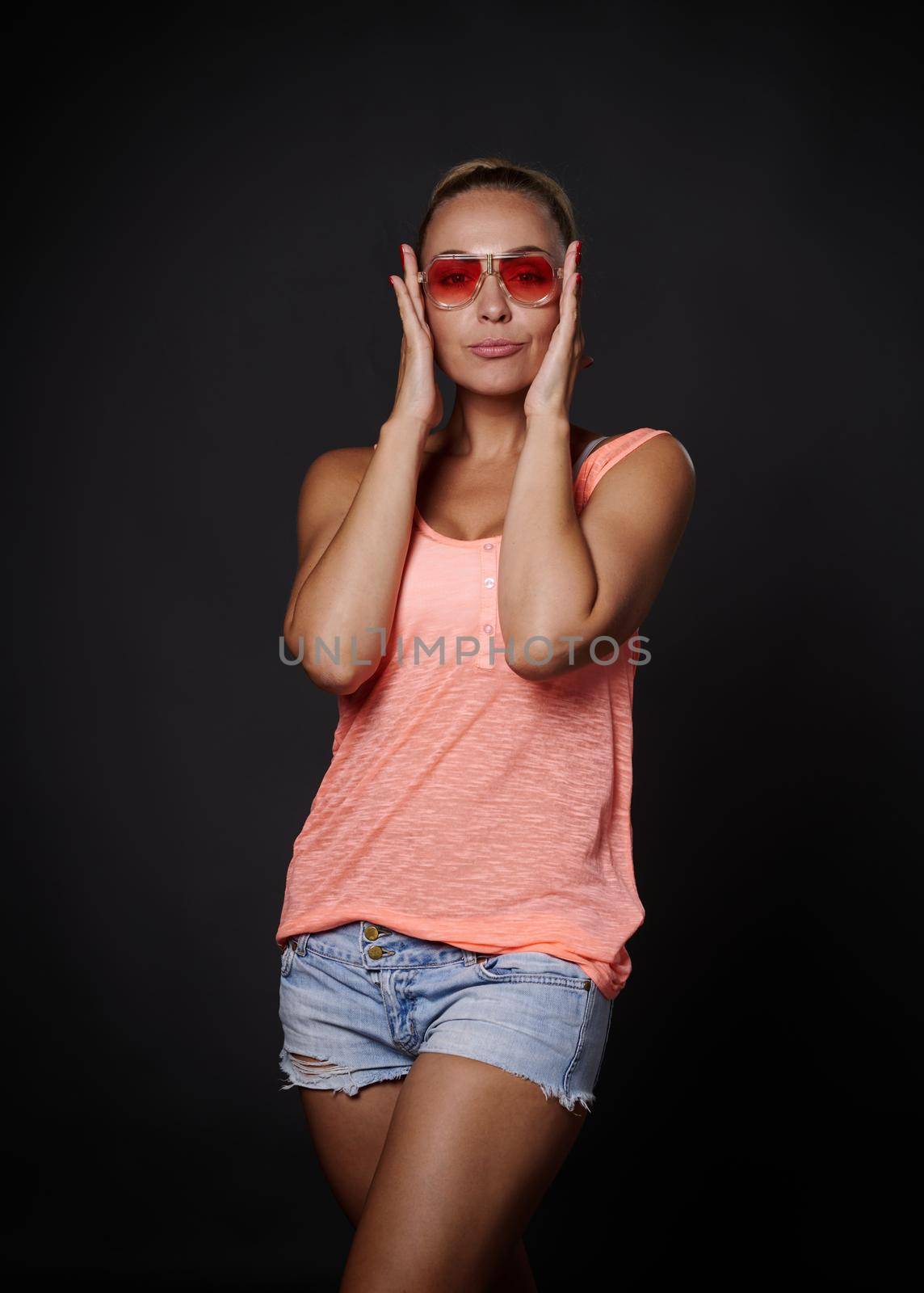Beautiful blonde middle aged Caucasian woman in casual summer clothes looking through her pink sunglasses and showing tongue to the camera, posing over black background with copy space
