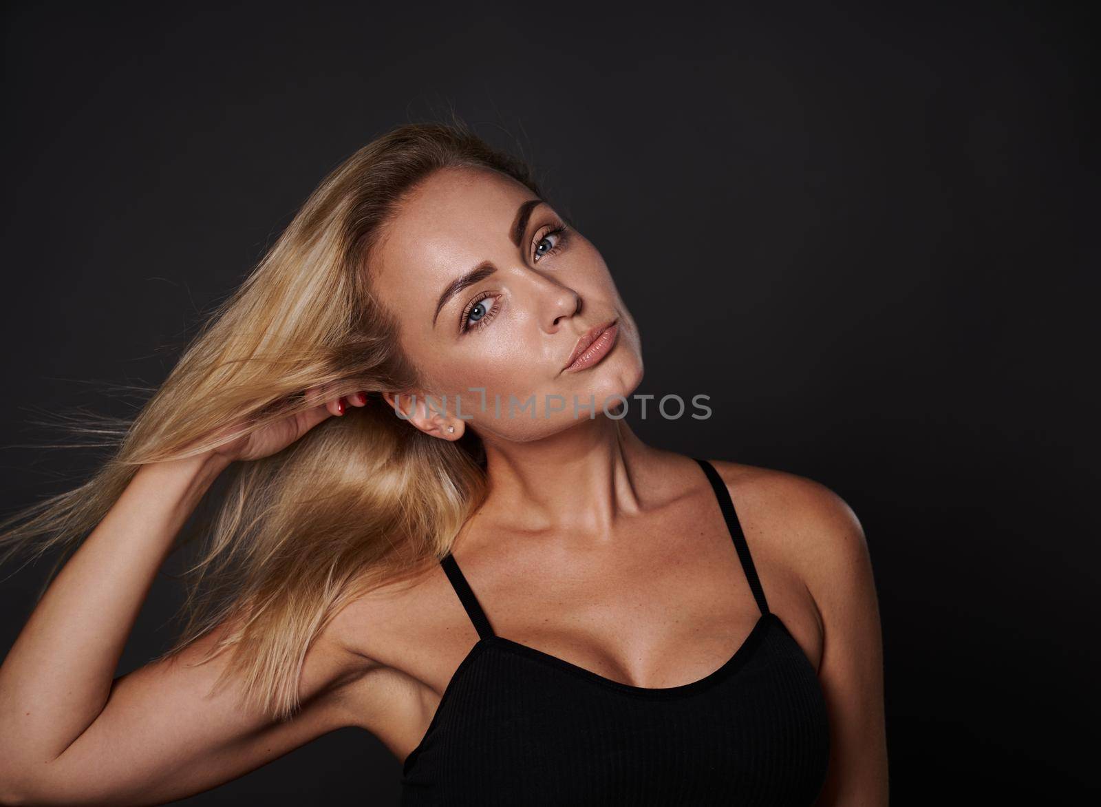Close up fashion portrait of beautiful European woman with toned body, fresh clean tanned skin touching her shining blond hair and looking at camera on black background by artgf