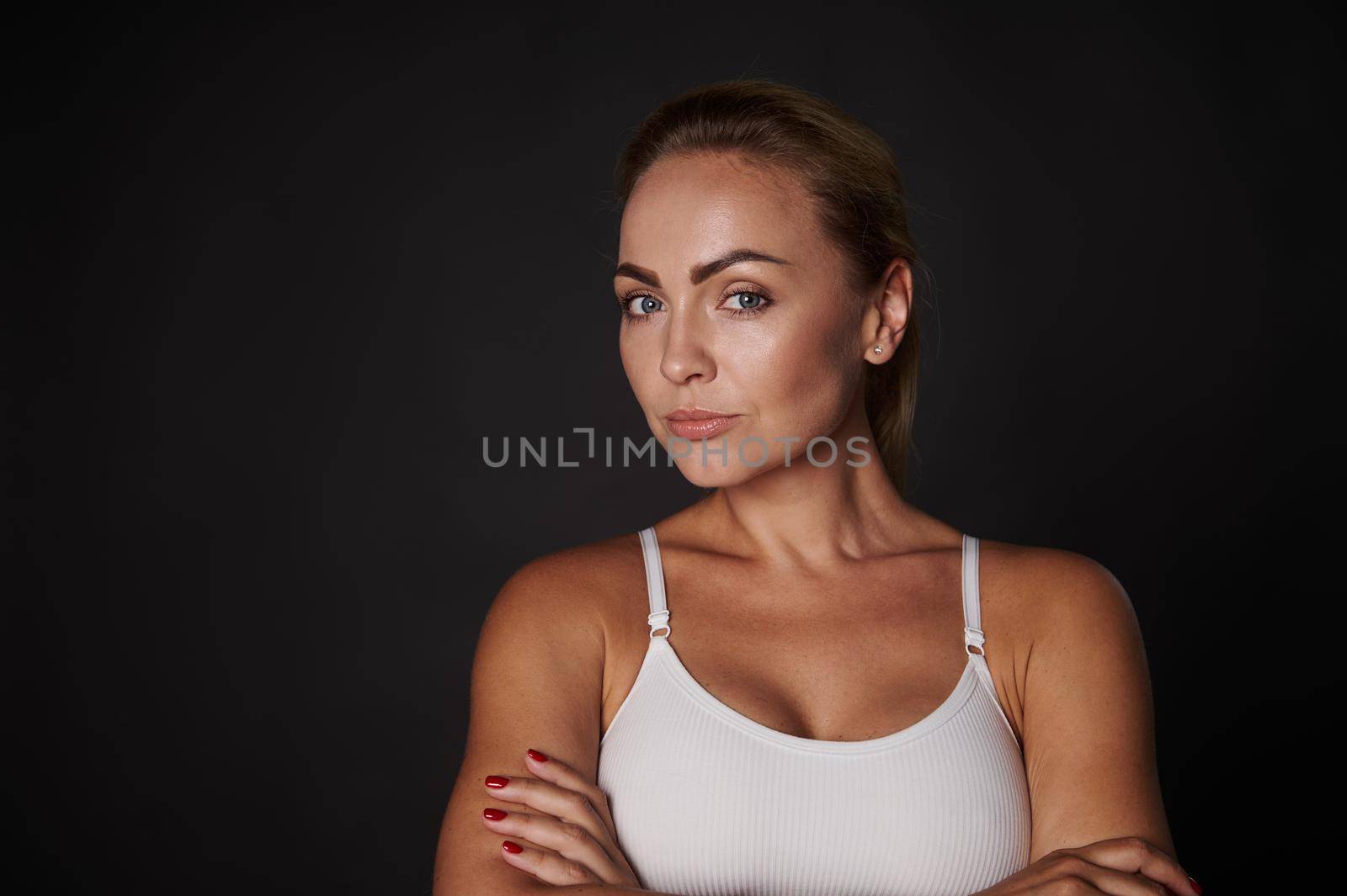 Waist length portrait of an attractive young blonde woman with natural makeup and beautiful aesthetic body wearing a white top standing with crossed arms against black background with copy space
