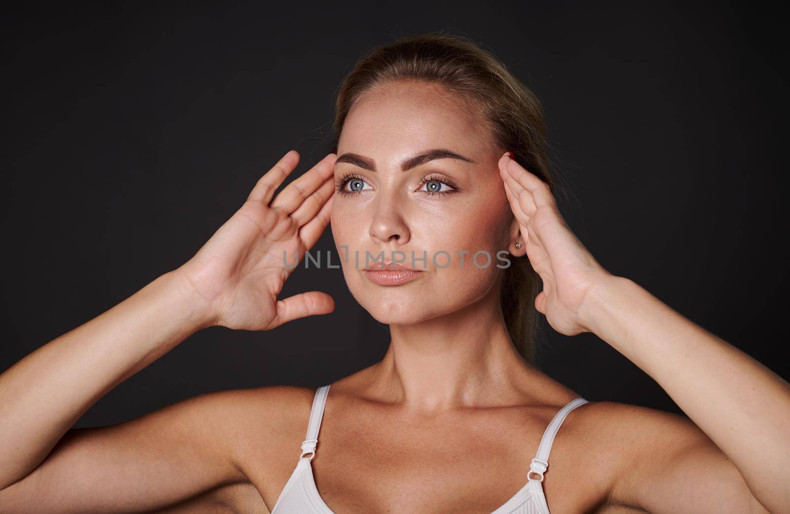Close-up portrait of an European pretty woman with fresh glowing skin holding hands on her temples, doing massage movements on her face. Anti-aging concept, smoothing, rejuvenating beauty treatment by artgf