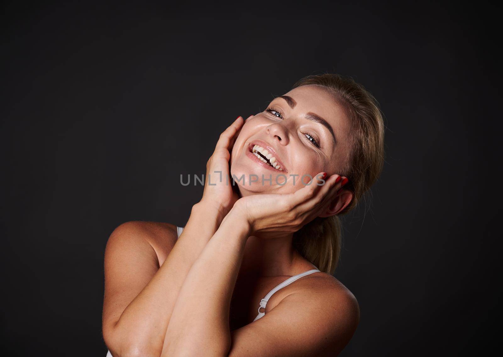 Attractive Caucasian middle aged woman with fresh glowing skin smiles with beautiful toothy smile, isolated over black background. Anti-aging concept, smoothing, rejuvenating beauty treatment by artgf