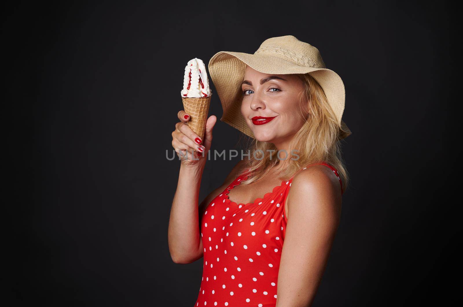 Attractive blonde woman in red swimsuit with white polka dots, holding delicious ice cream cone , smiling beautiful toothy smile, posing on black background copy space by artgf