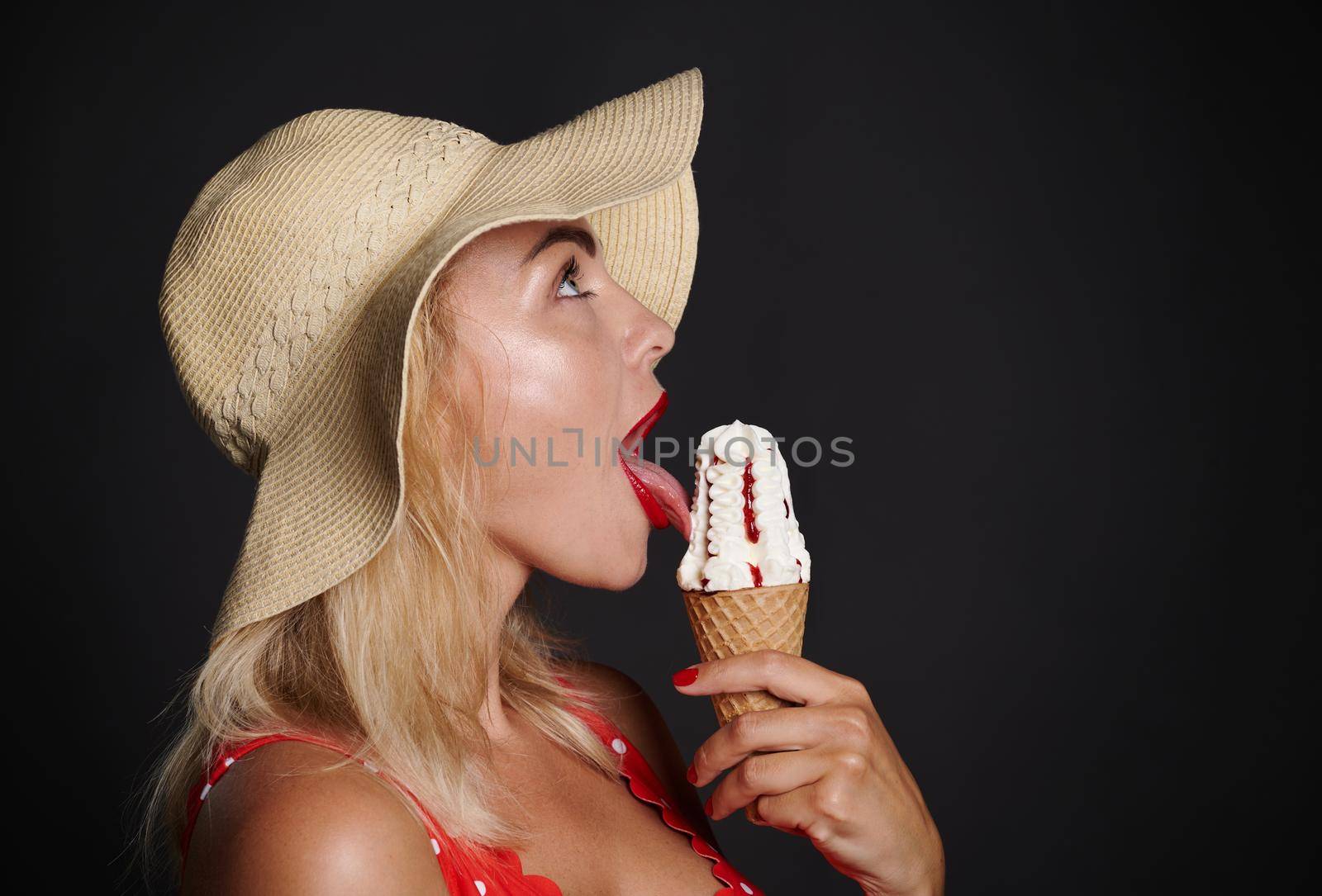 Close-up pretty blonde European woman in summer straw hat and red swimsuit with white polka dots tasting a delicious strawberry ice cream cone posing against black background with copy space for ads