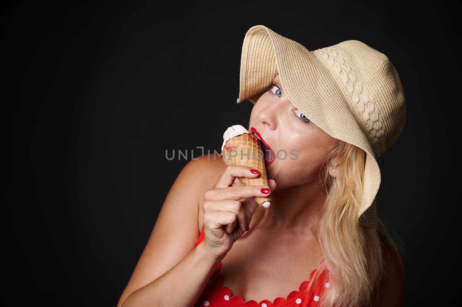 Attractive European blonde woman in summer hat holding a delicious ice-cream in her hands and smiles with beautiful toothy smile looking at camera, isolated over black background with copy ad space by artgf