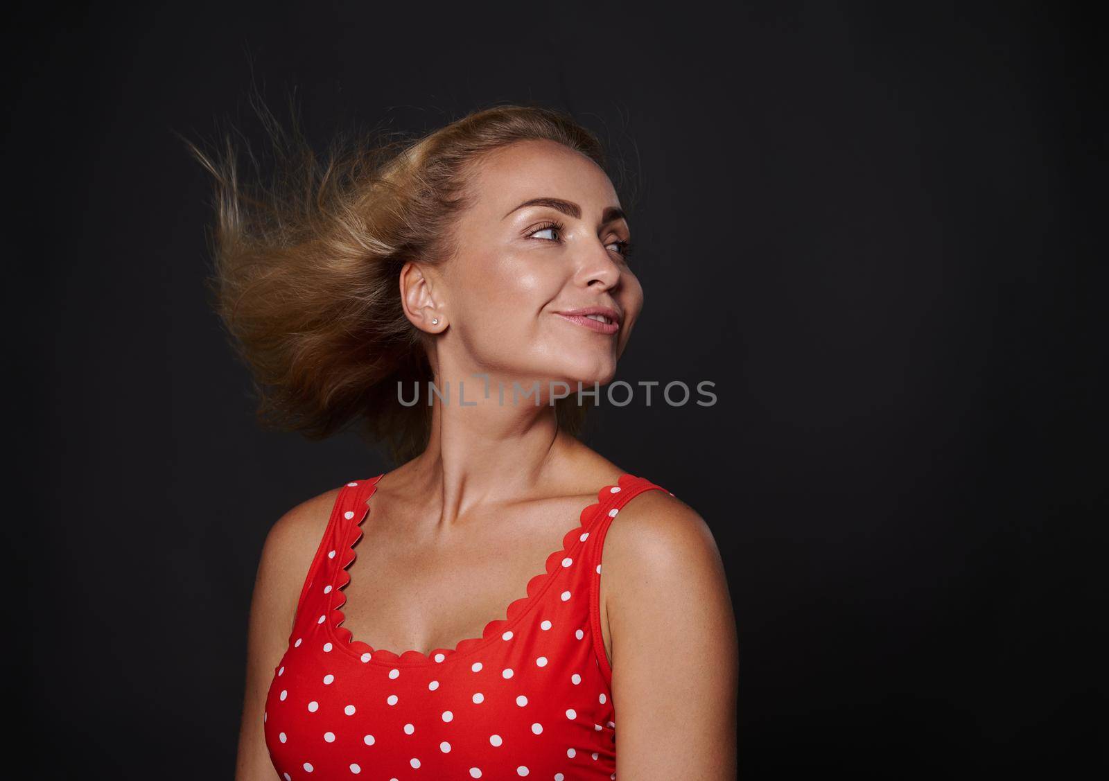 Smiling attractive stunning blonde Caucasian woman with natural makeup and sun tanned skin wearing red swimsuit with polka dots and looking aside at a copy space on black background. Summer concept