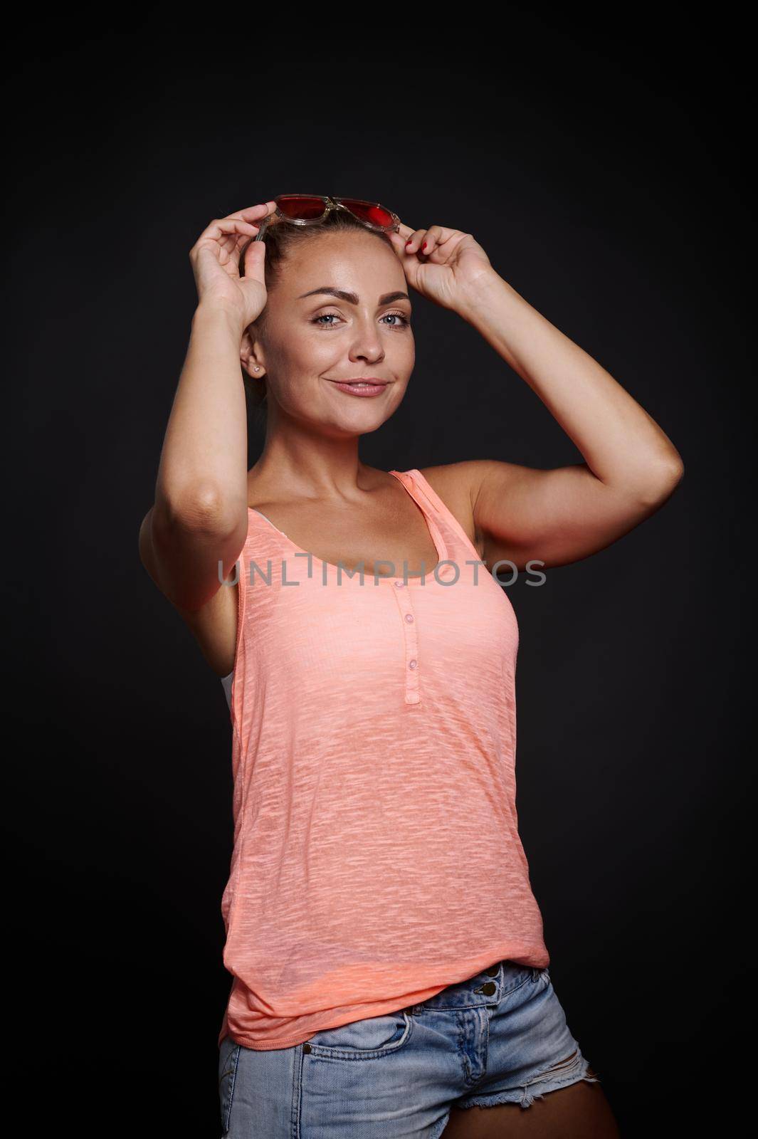 Attractive blonde Caucasian woman with tanned skin and beautiful fit body holding her hands near her face and confidently looking at camera against black background with copy ad space