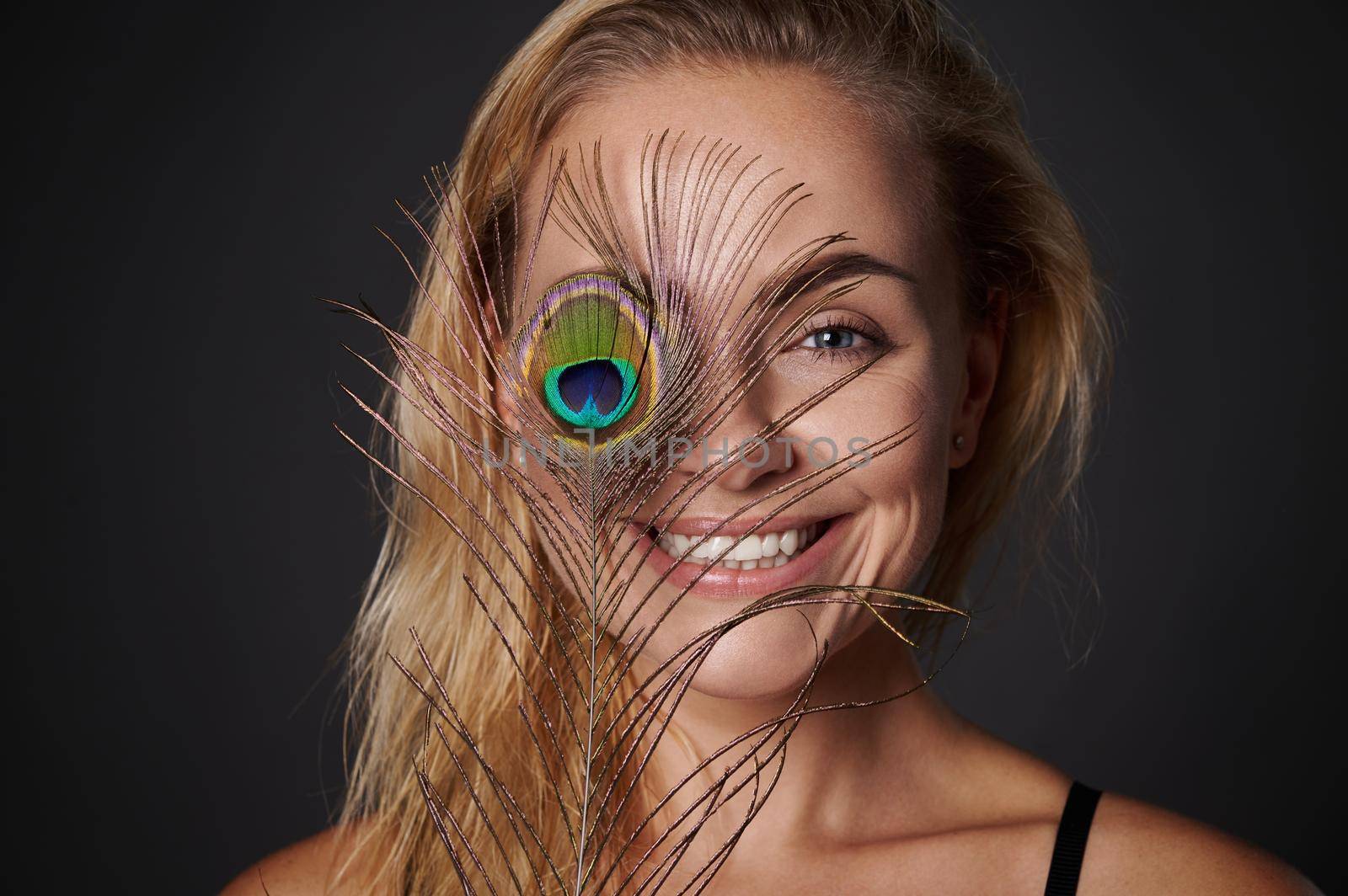 woman, with beautiful healthy glowing skin holding a peacock feather near her face. Black background copy space. by artgf