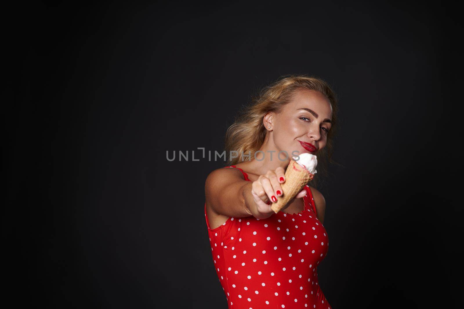 Charming stunning beautiful blonde Caucasian young woman wearing red swimsuit and holding ice-cream cone in outstretched hand, showing it to camera, isolated over black background with copy ad space by artgf