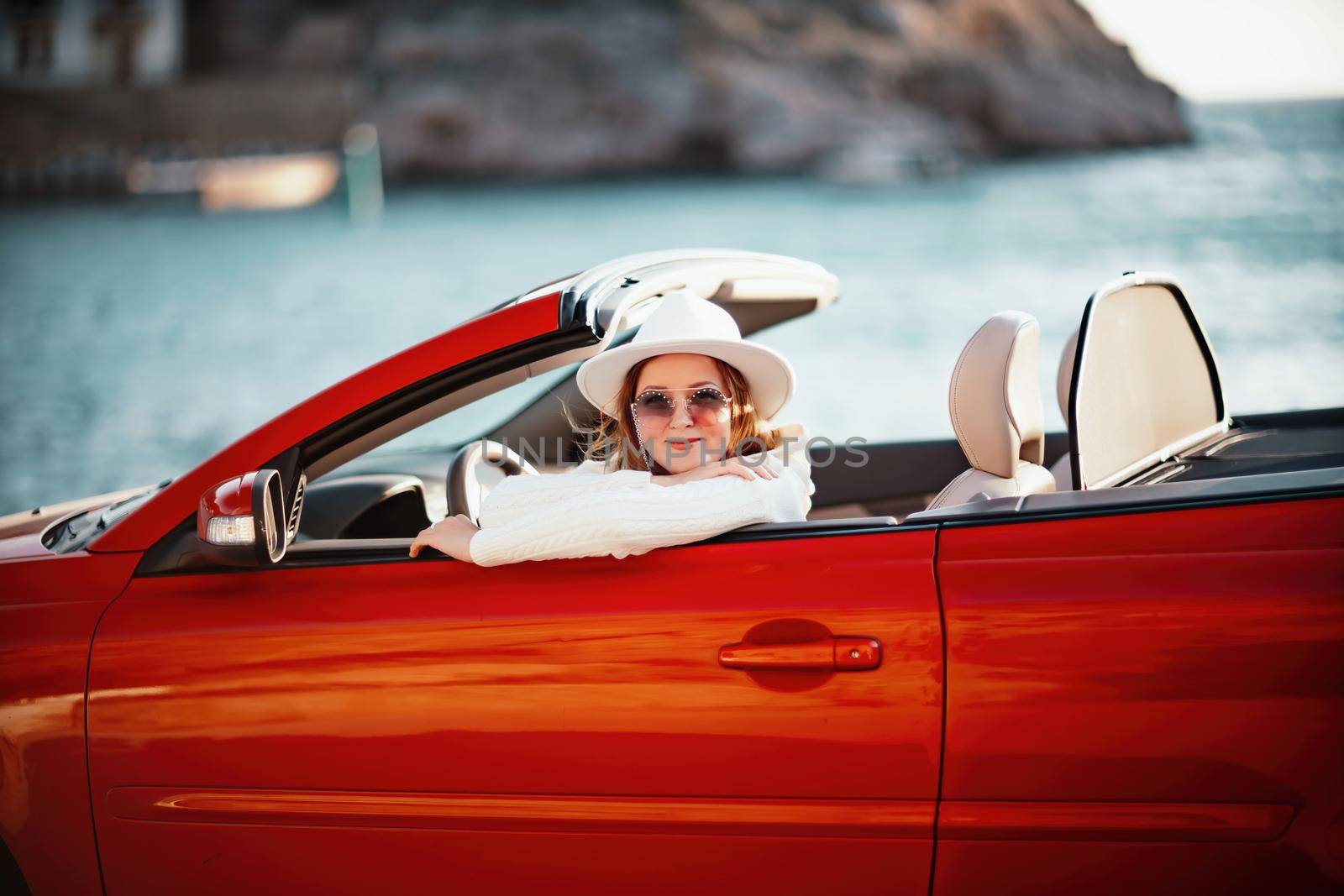 Outdoor summer portrait of stylish blonde woman driving red car convertible. Fashionable attractive woman with blond hair in a white hat in a red car. Sunny bright colors taken outdoors against the sea. by Matiunina