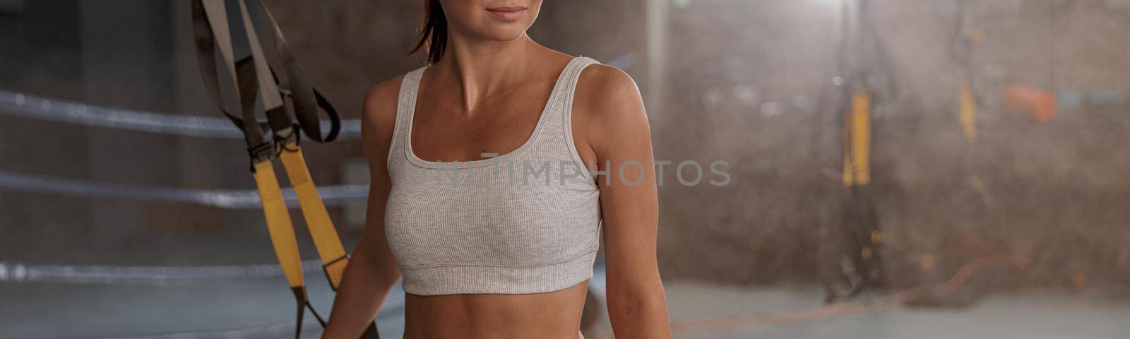 Beautiful slim body woman with flat belly looking to side while standing with suspension straps at gym club