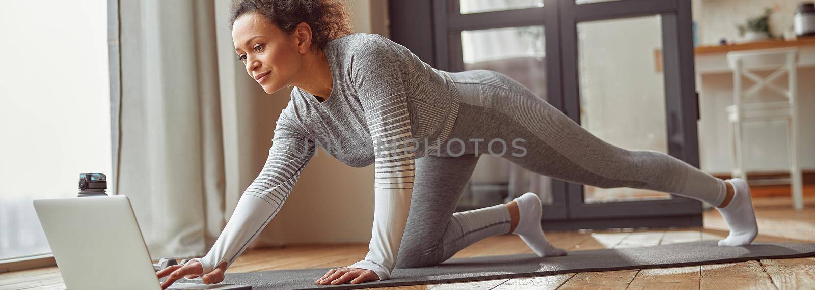 Low angle of smiling female in sportswear doing training on mat with video guide on laptop