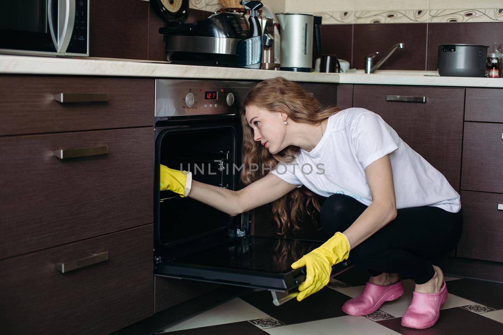 View of young woman in yellow protective gloves with rag cleaning oven at home kitchen. Brunette girl with curly hair washing oven on kitchen holding hand inside. Housework concept.
