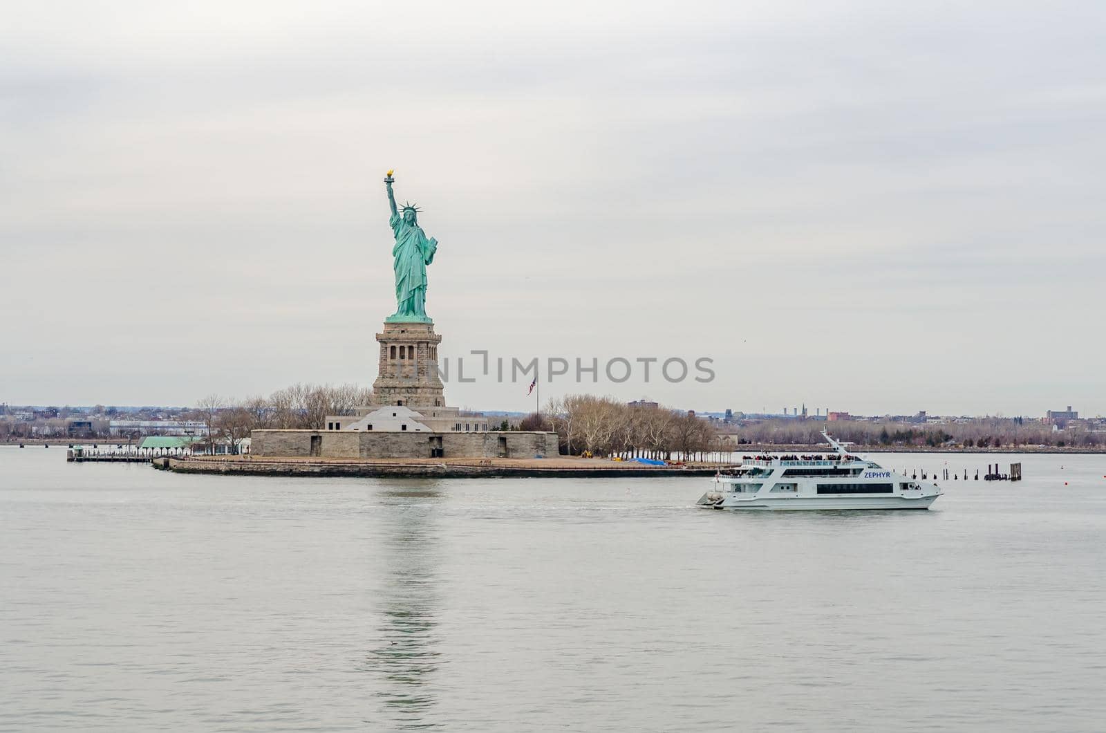 Statue of Liberty National Park, New York City with white ZEPHYR Ferry with lots of tourists on deck passing by in the forefront, during winter day with overcast, horizontal