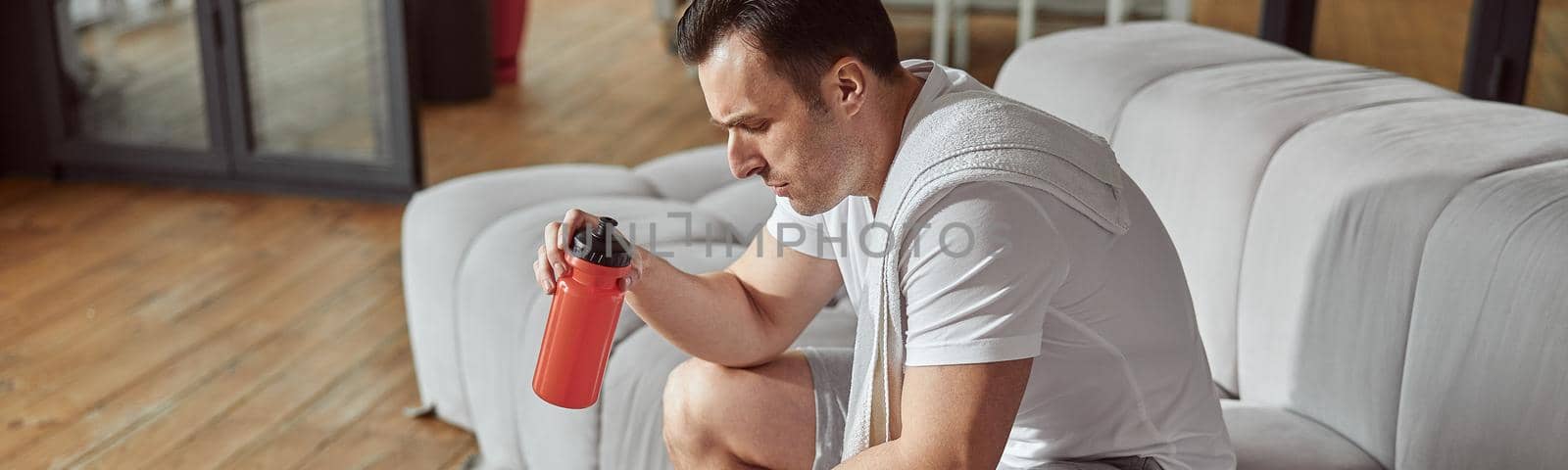 Top view of thirsty man with botle of water and towel sitting on sofa after workout at home