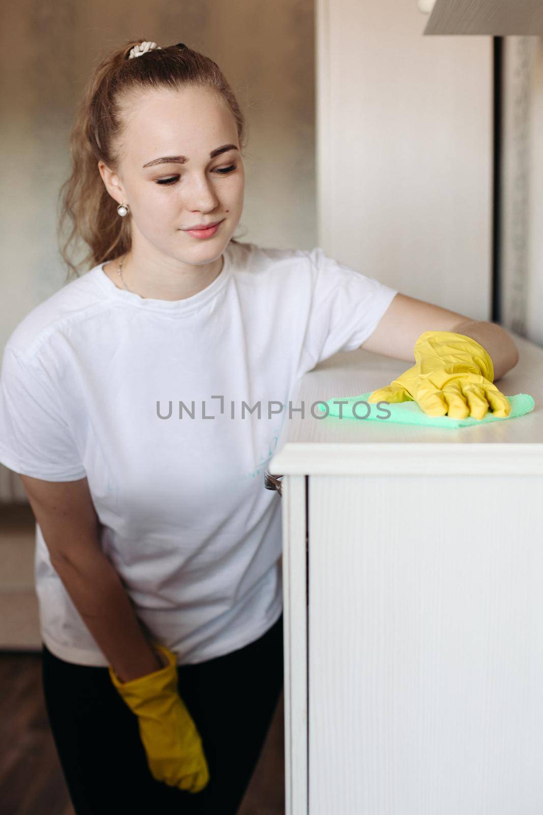 Pretty girl wearing in yellow protective rubber gloves, wiping furniture with special mint rattling. Employee of cleaning company wearing in uniform working at home client. Housecleaning concept.