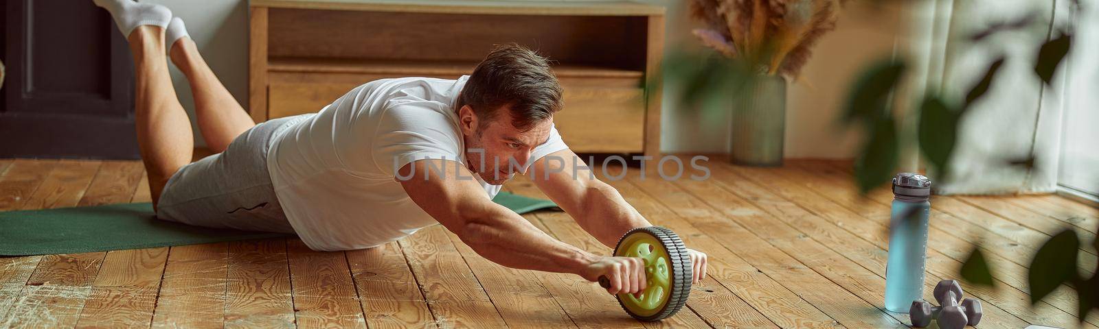 Sporty guy is using abs wheel in living room while watching online workout on notebook