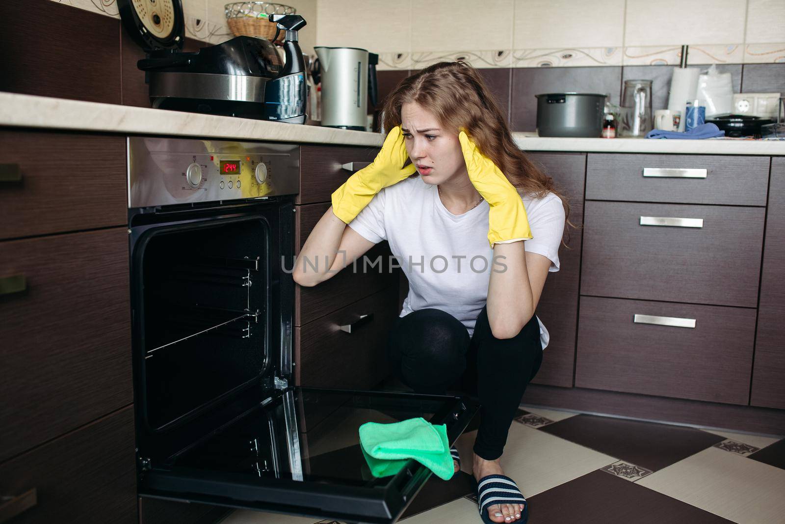 Young woman in yellow protective gloves holding head by hands, looking dirty oven at home kitchen. Brunette girl with curly hair washing oven on kitchen holding hand inside. Housework concept.