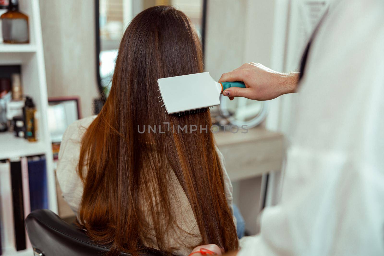 Hand of hairdresser brushing long and shiny brown hair of young woman at beauty salon by Yaroslav_astakhov