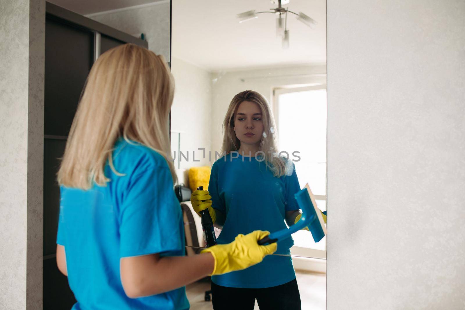 Female employee of cleaning company wearing in uniform working at client home. Blonde girl wearing in yellow protective rubber gloves, with special detergent spray and brush, washing mirror.