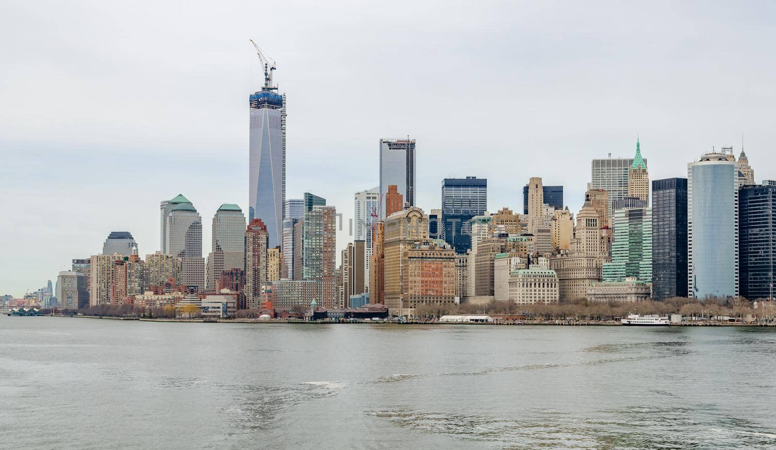 Manhattan Skyline with One World Trade Center with construction area, Crane on top of it and Hudson river in forefront, during winter day with overcast, horizontal