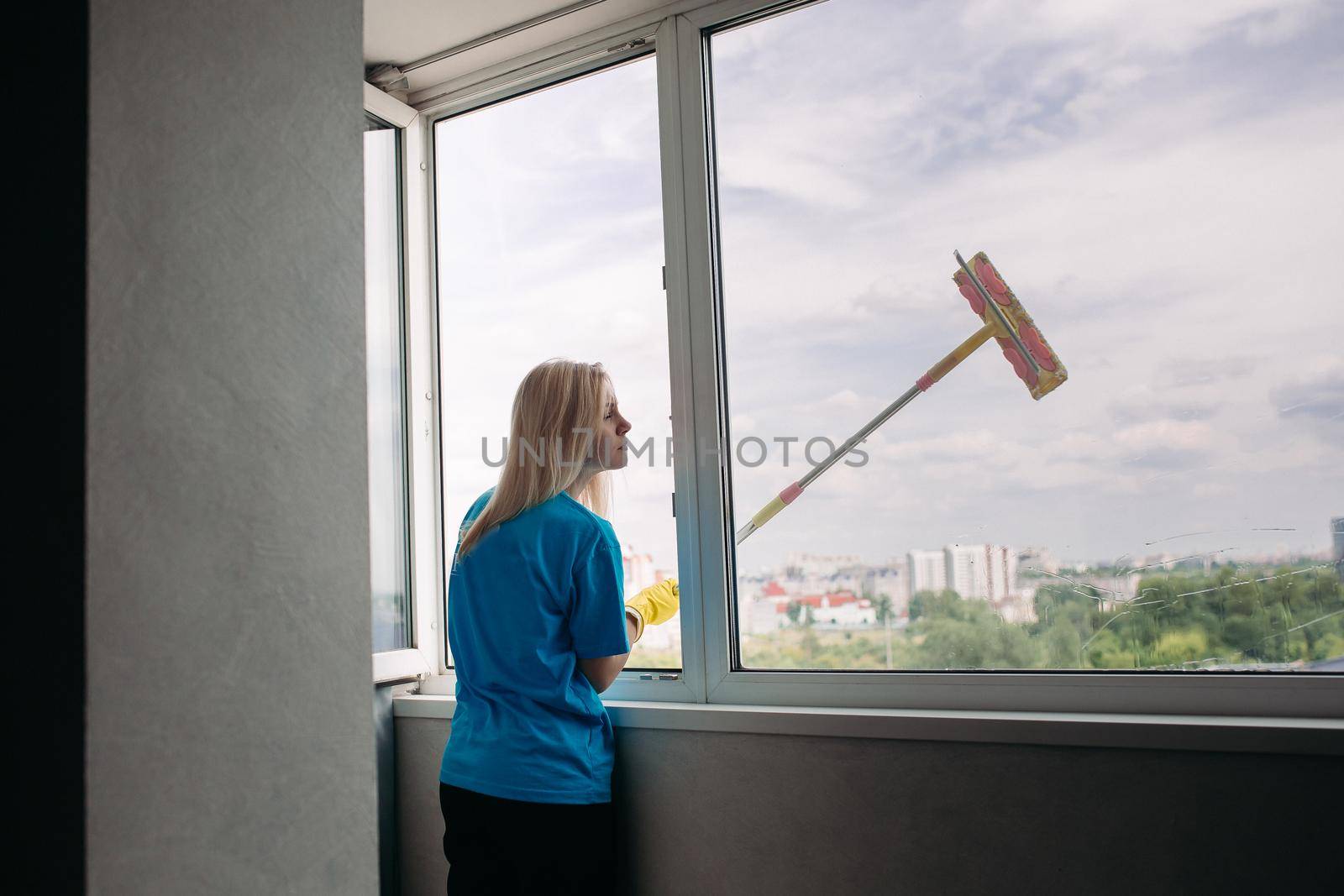 View of young blonde girl wearing in blue shirt and yellow protective gloves with special brush, cleaning large open window. Female employee of cleaning company wearing in uniform working alone.