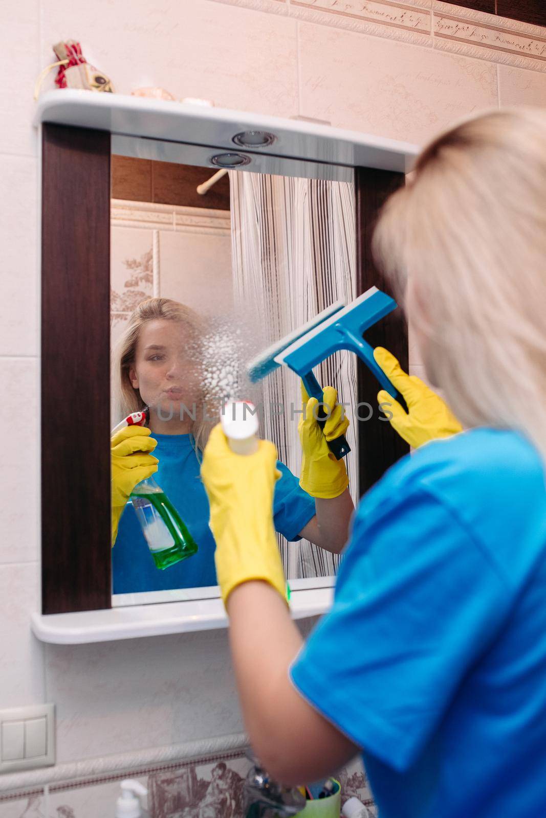 Happy young woman cleaning with spray and using squeegee to wash mirror at bathroom. Cute blonde girl wearing in yellow protective glove, working at home. Housework and housekeeping concept.