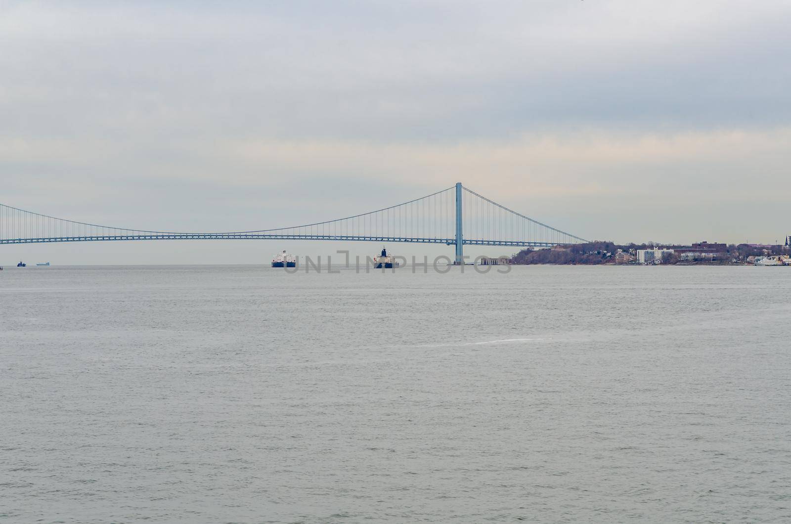 Two industrial ships underneath blue Verrazano-Narrows Bridge, New York City, at the horizon, during winter evening with overcast, horizontal
