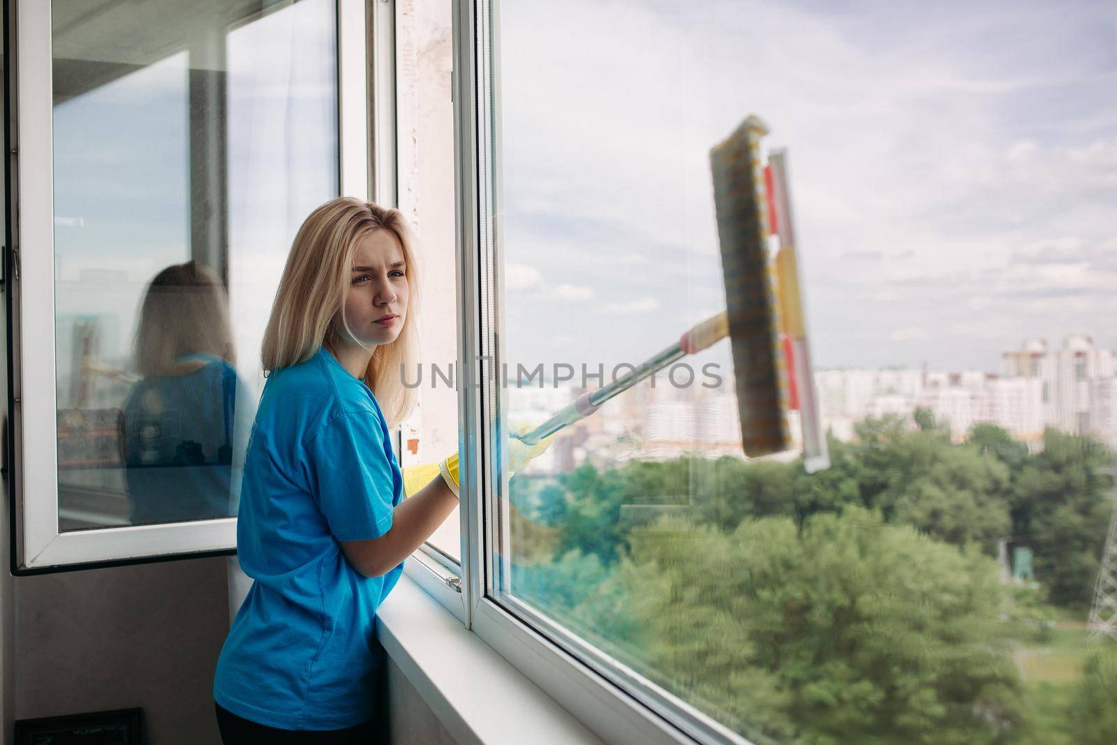 Young girl wearing in yellow protective rubber gloves with special brush, cleaning large open window overlooking park. Female employee of cleaning company wearing in uniform working at client home.